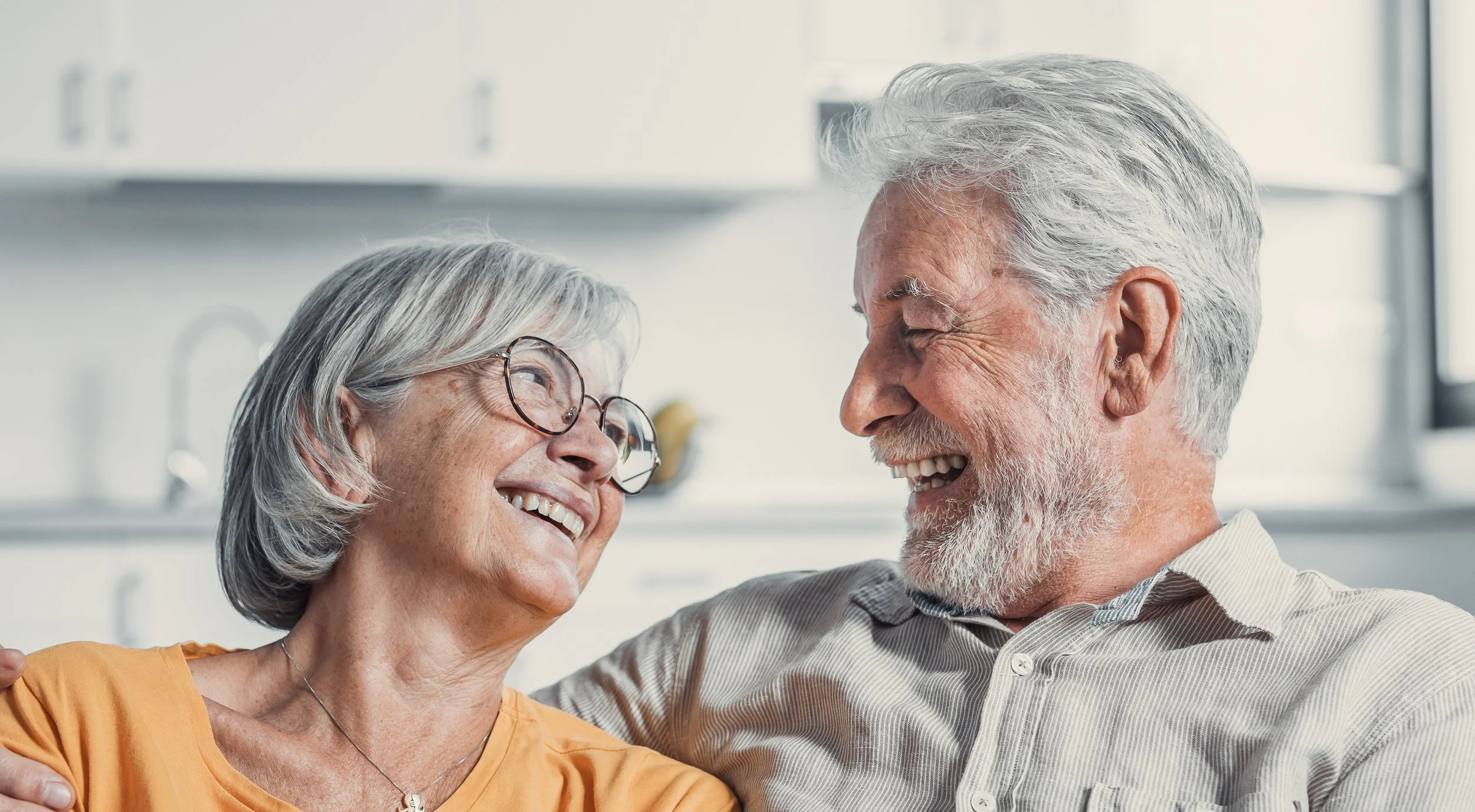 An elderly couple shares a joyful moment, smiling and looking into each other's eyes, in a bright, modern kitchen.
