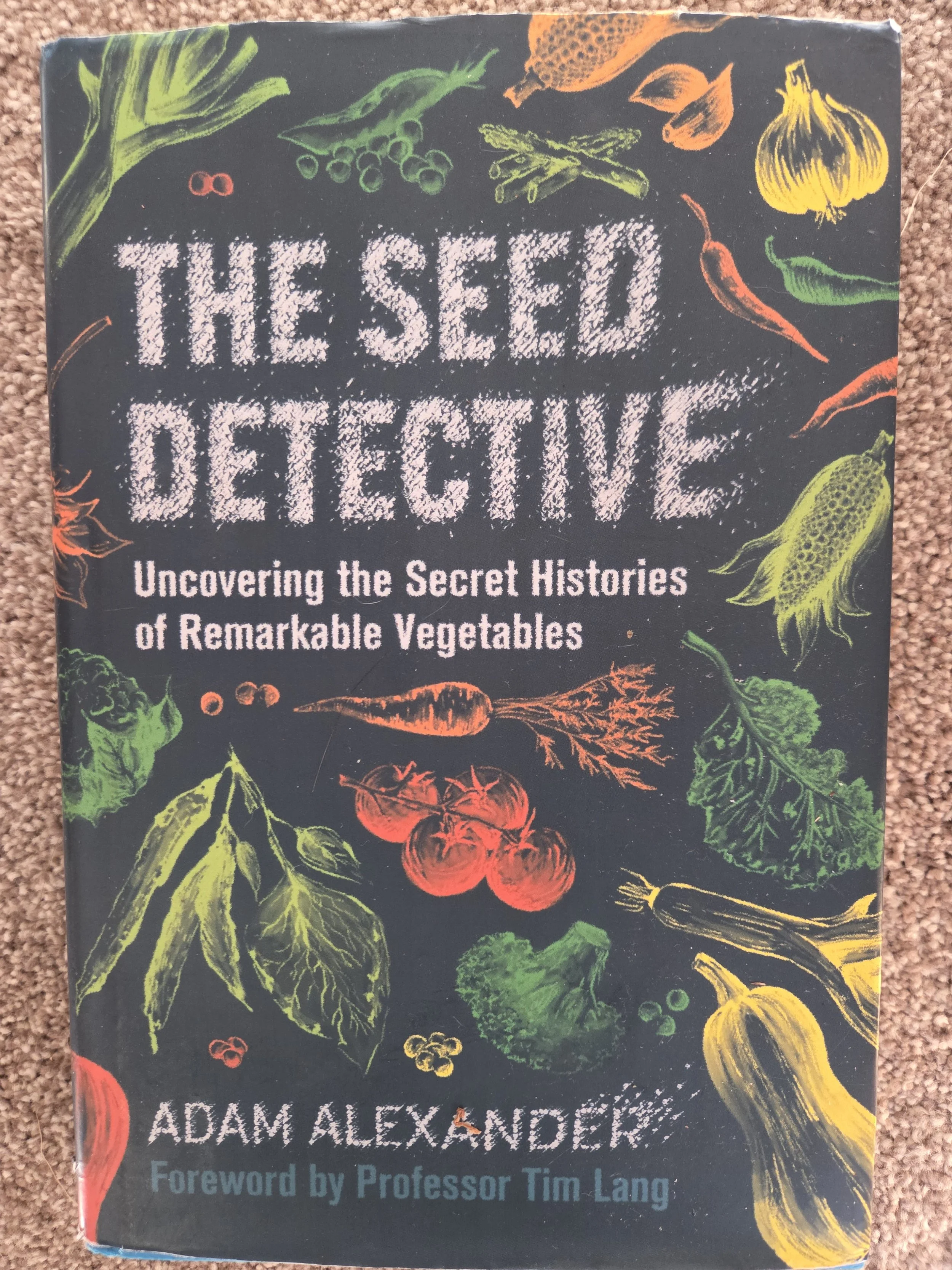 Cover of a book titled 'The Seed Detective: Uncovering the Secret Histories of Remarkable Vegetables' by Adam Alexander with a foreword by Professor Tim Lang. The cover features colorful sketches of various vegetables such as tomatoes, carrots, lettuce, broccoli, garlic, onion, peppers, and zucchini on a black background.