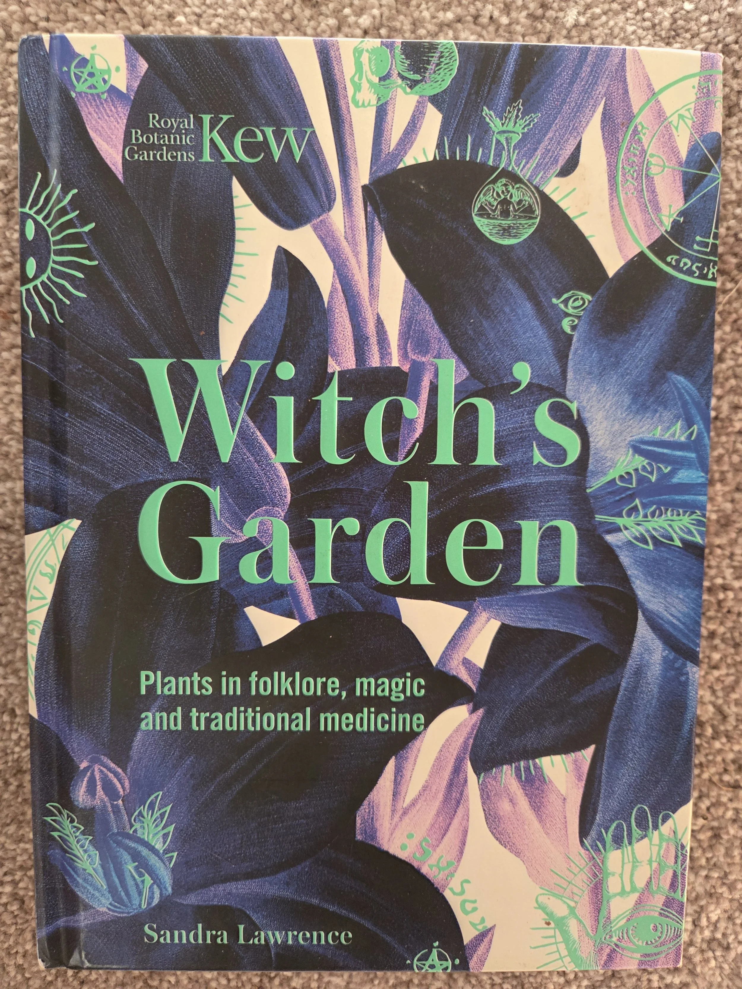 Book cover titled 'Witch's Garden' by Sandra Lawrence, featuring illustrations of dark blue leaves and plant motifs with green overlay symbols, including an eye, a sun, and a celestial map, from the Royal Botanic Gardens Kew.
