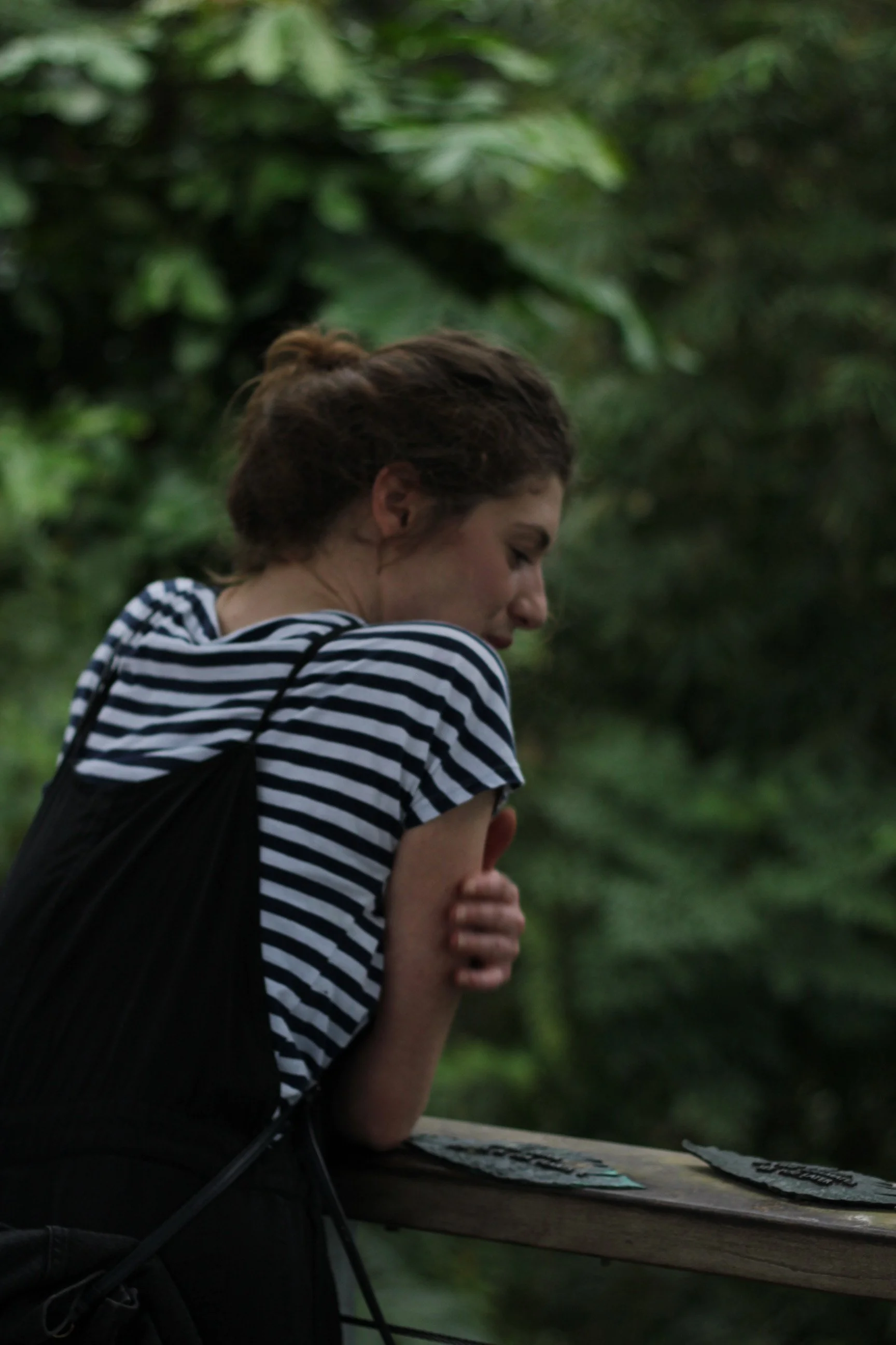 A woman with brown hair, wearing a black and white striped shirt, leaning on a wooden railing outdoors surrounded by lush green foliage.