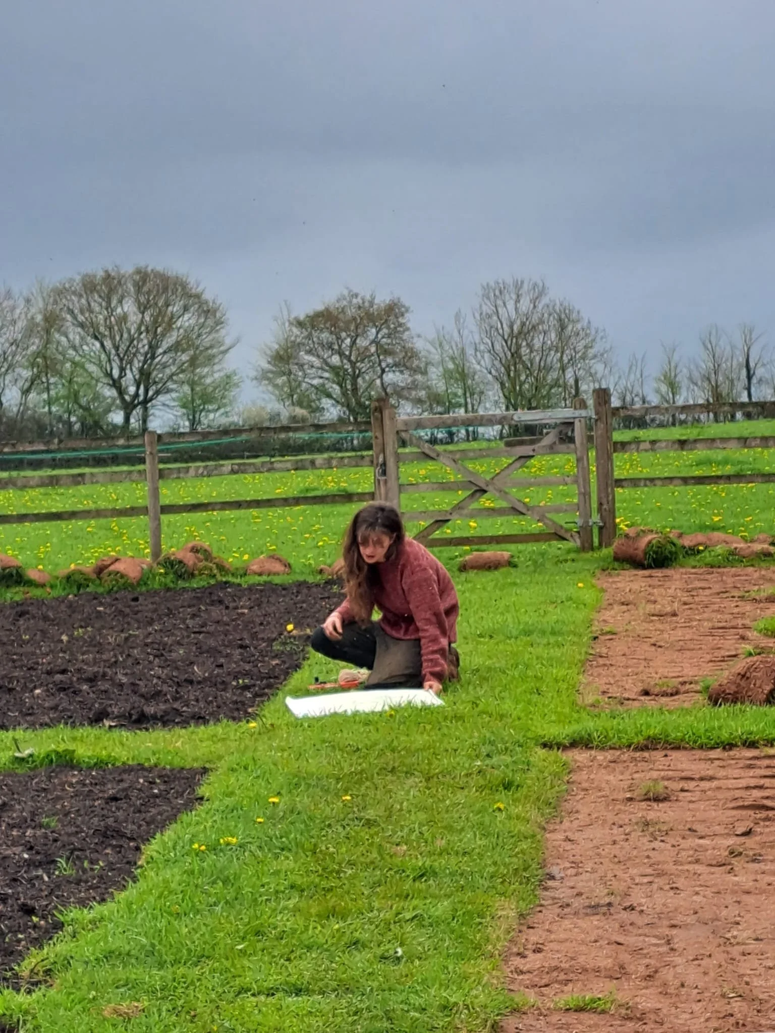 A woman kneeling on the grass near a garden bed, with a clipboard or notebook in front of her, on a farm or rural property with a wooden gate and trees in the background.