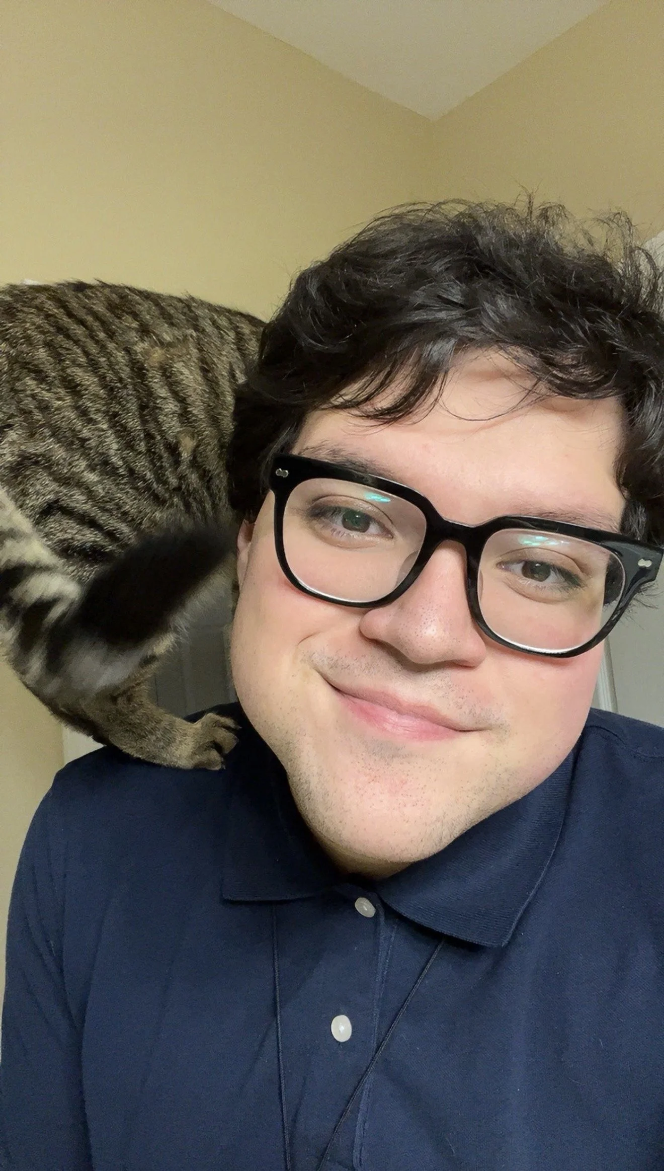A young man with glasses smiling, with a tabby cat perched on his shoulder, in a room with beige walls.