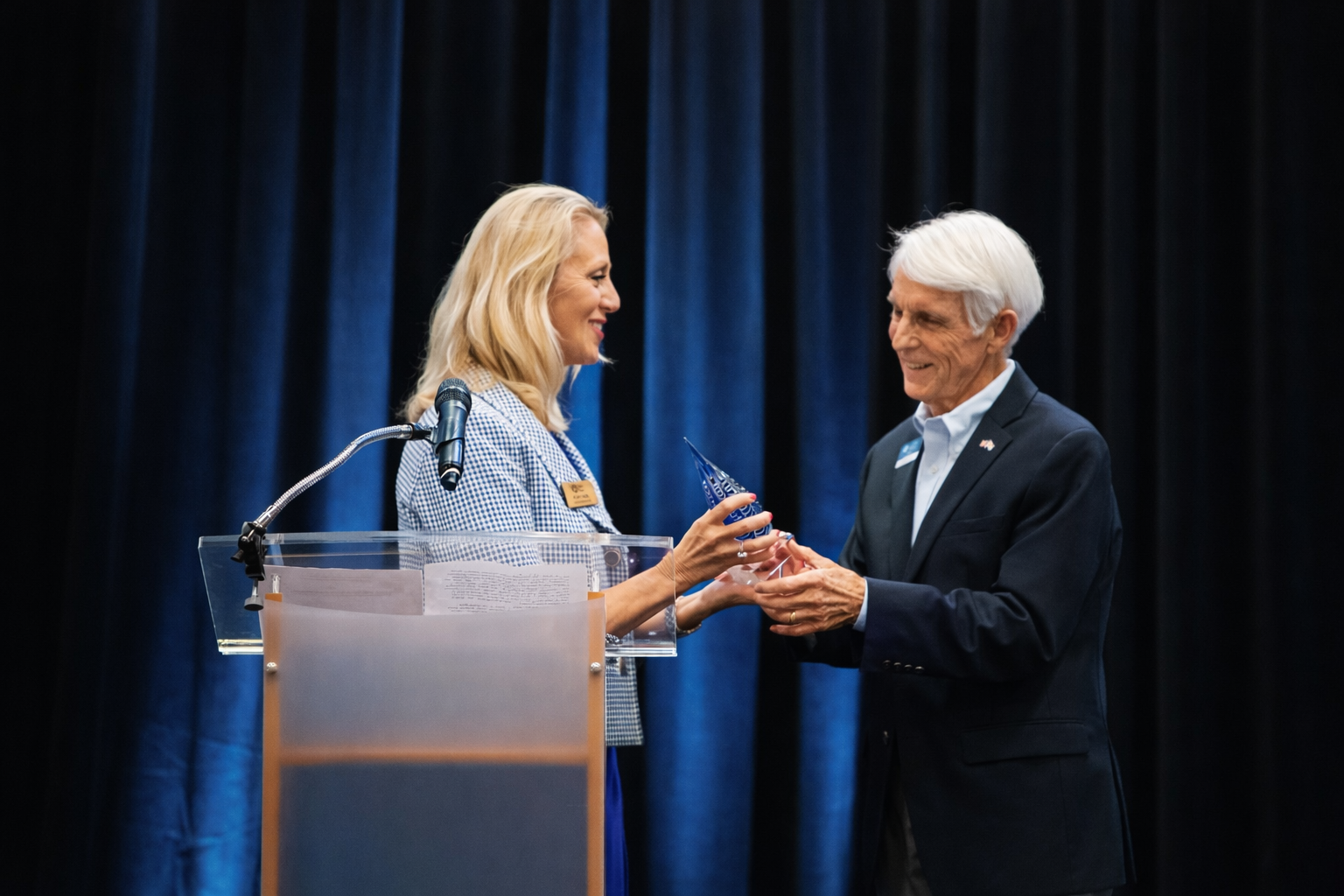 A woman presenting an award to a man on stage with a blue curtain background.