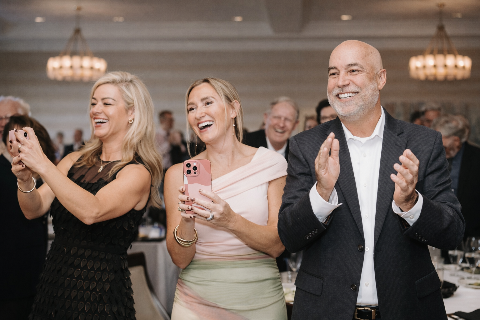 People smiling and clapping at a formal event, with two women holding phones, one man clapping, and others blurred in the background.