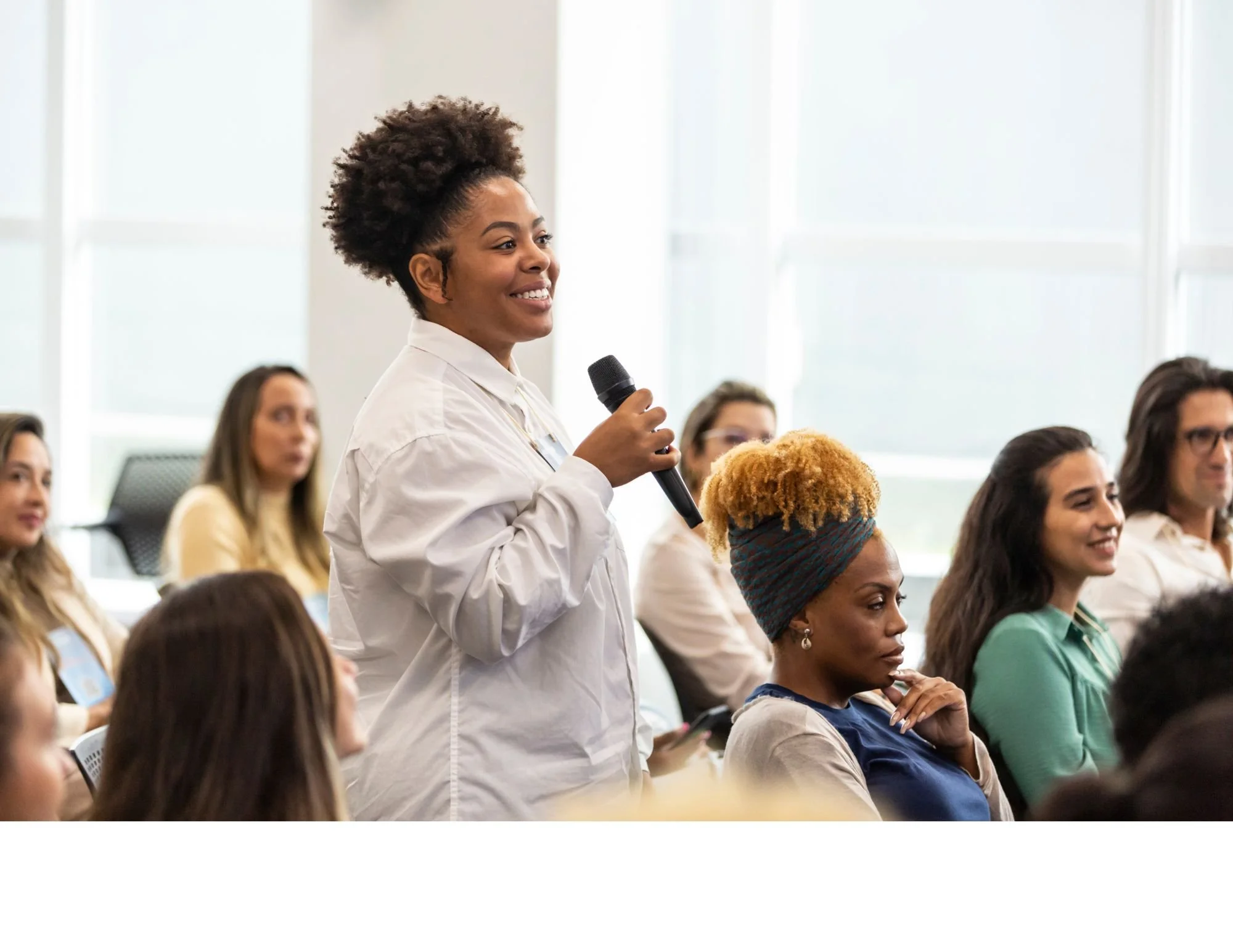 A woman with curly hair speaking into a microphone at a conference or seminar, surrounded by an audience of women seated in a bright, modern room.