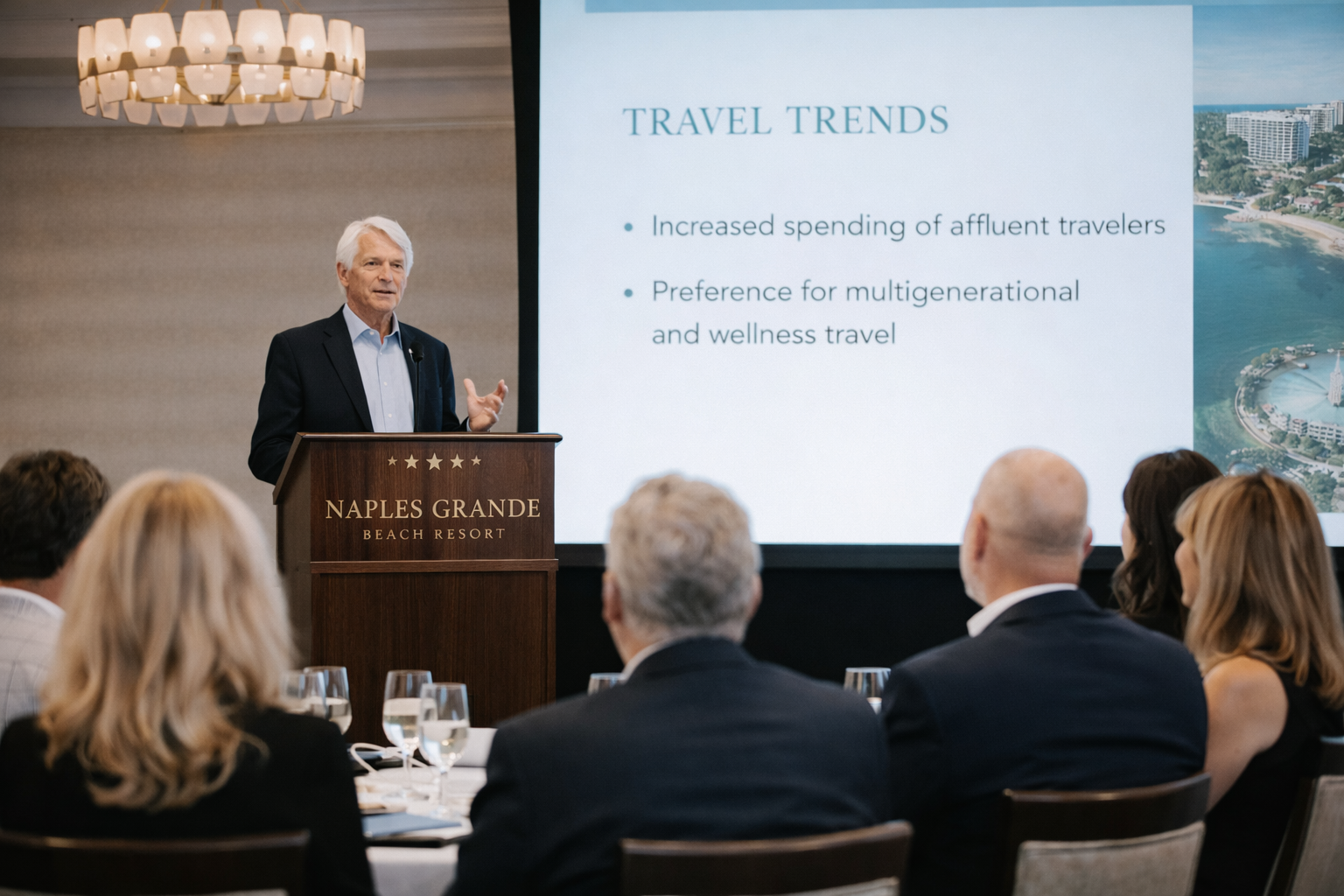 A man giving a presentation at a conference at Naples Grande Beach Resort, with a screen behind him displaying travel trends.