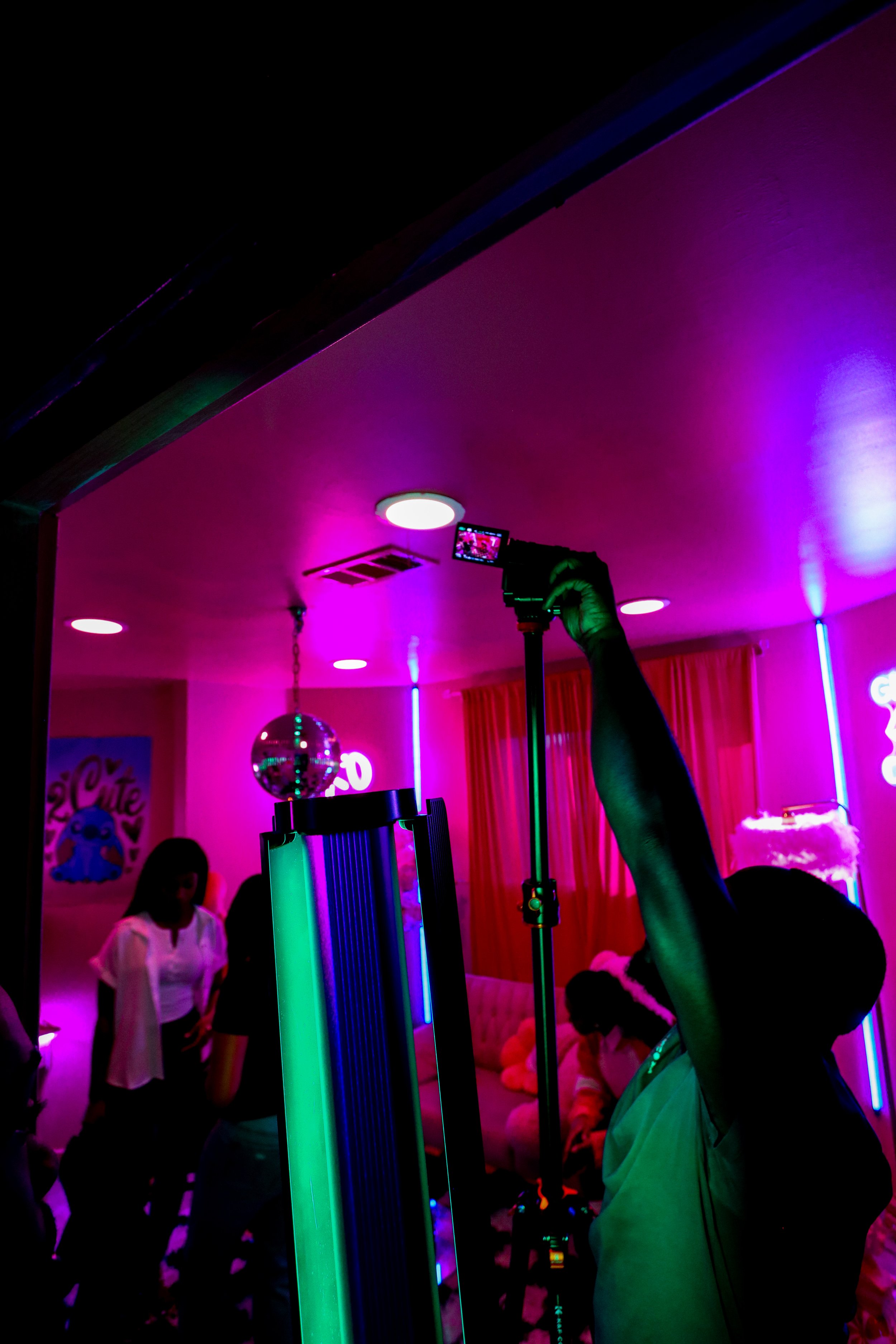 People at a colorful, neon-lit indoor party with pink and purple lighting, a disco ball, a woman taking a photo with her phone, and others socializing in the background.