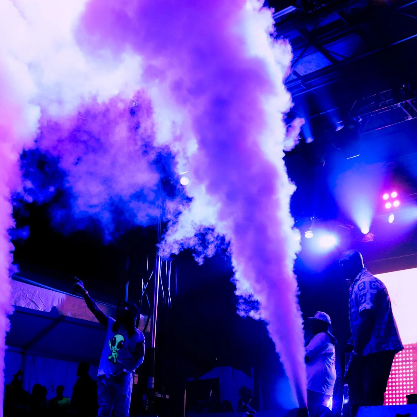 People on a stage with colorful fog, purple and blue lighting, and a large screen in the background at a concert or event.