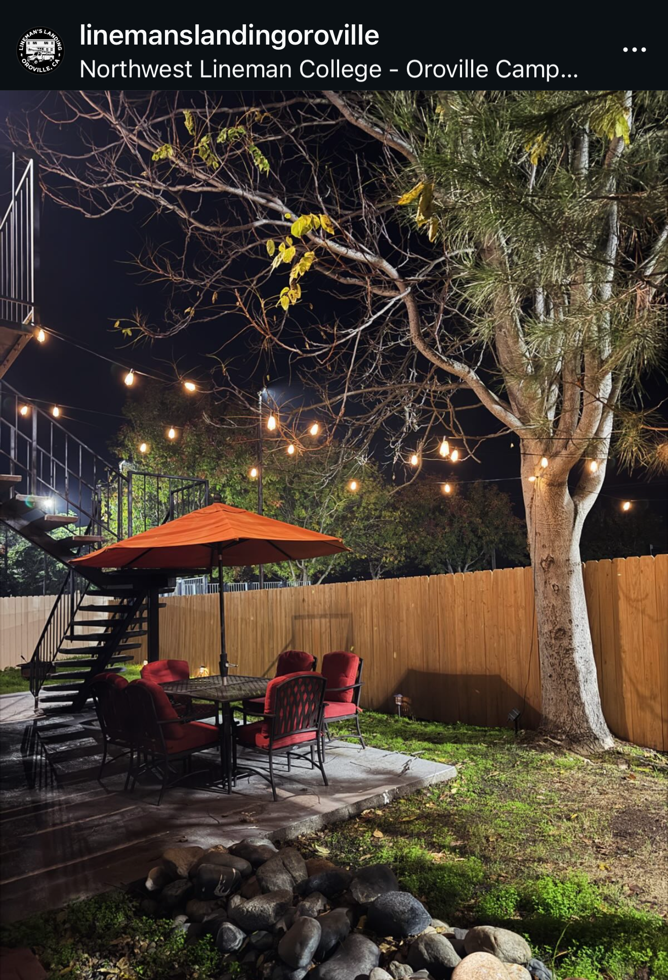 Nighttime scene of a backyard patio with a round table, six red cushioned chairs, an orange patio umbrella, string lights, a large tree, and a wooden privacy fence.