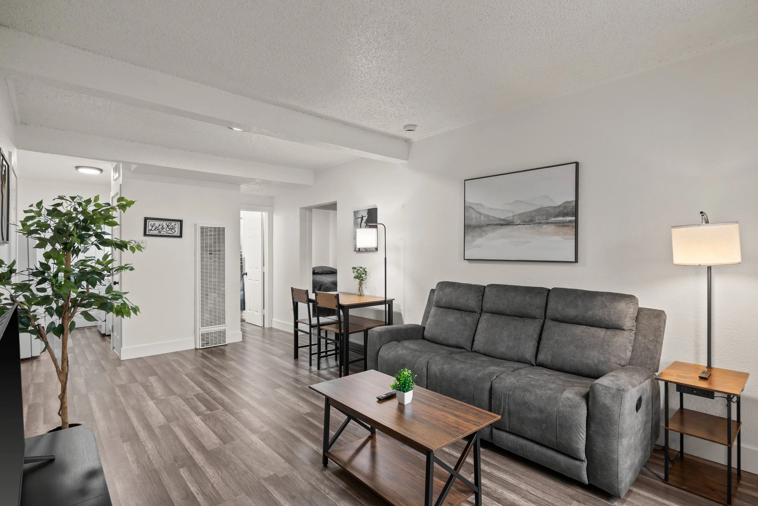 Modern living room with gray sofa, wooden coffee table, floor lamp, wall art, and dining area, with hardwood floors and white walls.