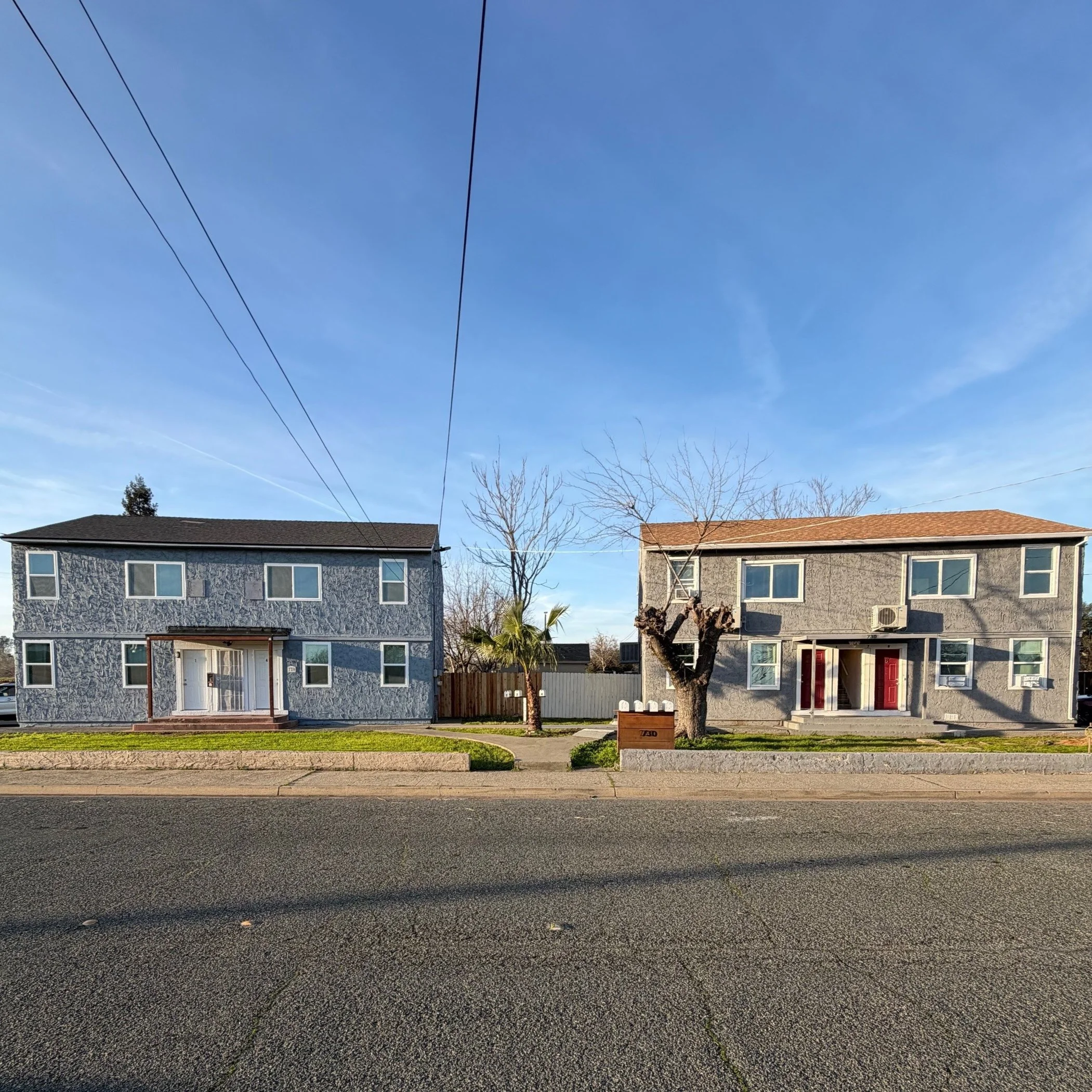Two residential buildings with trees and lawns in front, parked cars, and cables overhead on a clear day.