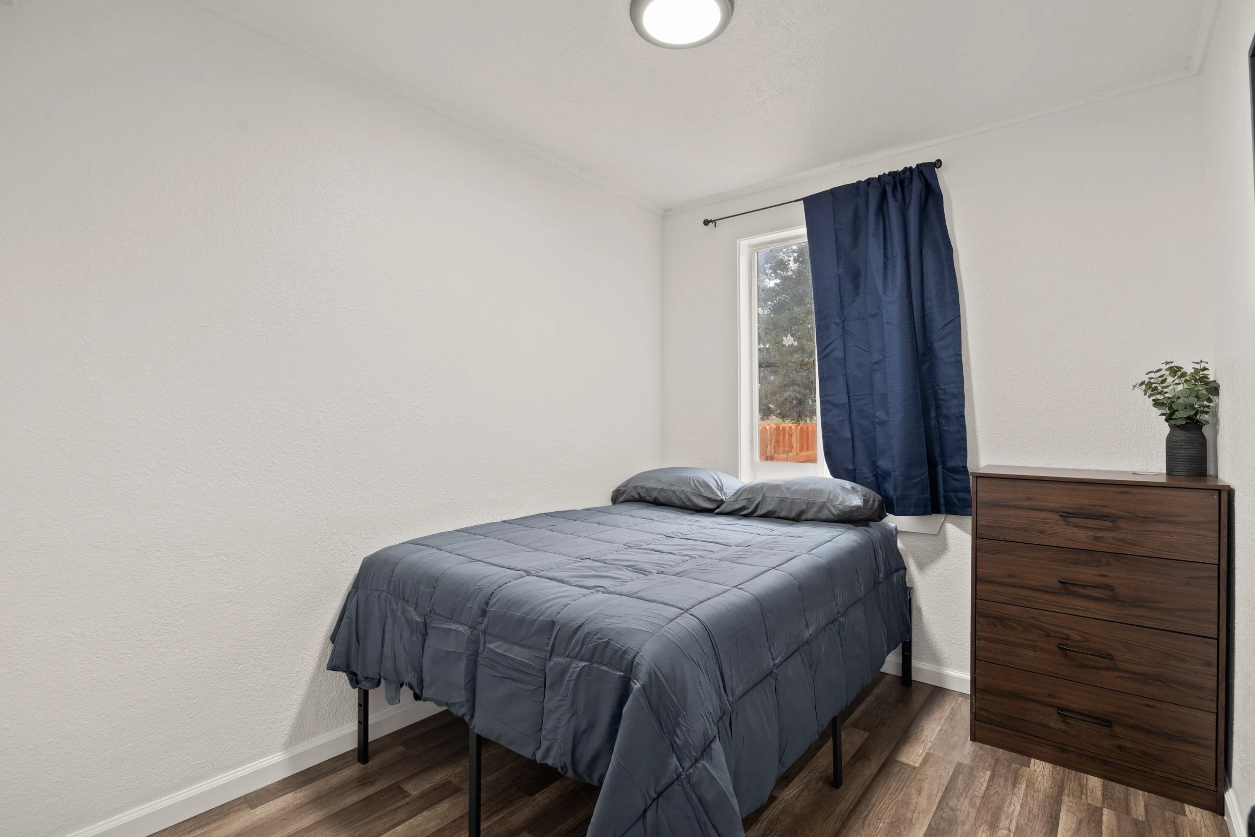 A minimalist bedroom with a bed covered in a dark gray comforter and two matching pillows, a dark wood dresser with a potted plant on top, white painted walls, a window with blue curtains, and dark wood flooring.