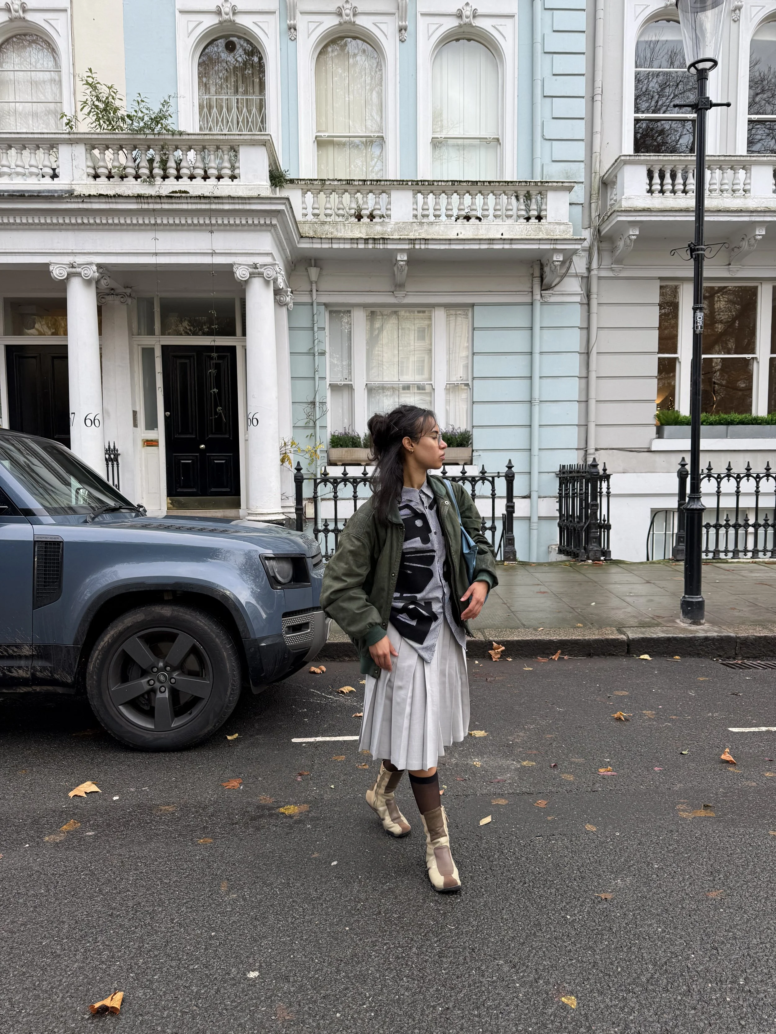 A woman walking on a city street with a grey car parked nearby. She is wearing a dark green jacket, a patterned shirt, a pleated grey skirt, and beige high-heeled boots. Behind her are pastel-colored Victorian-style houses with ornate details and black iron fences, and a black street lamp.