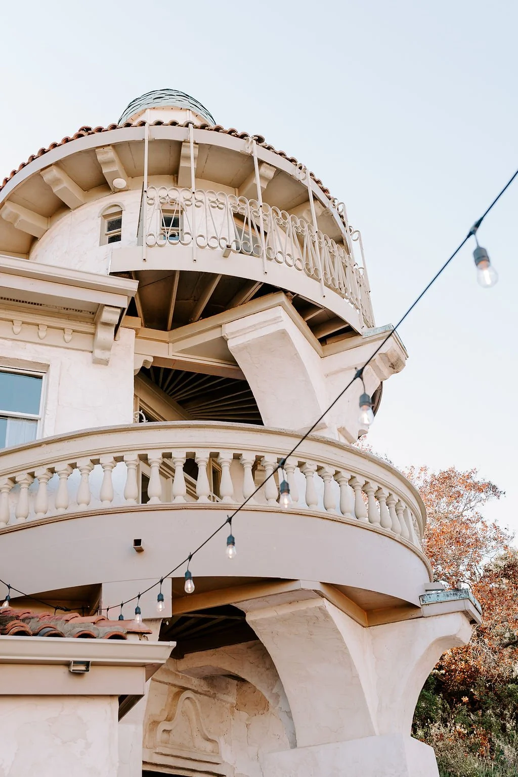 A white multi-story house with rounded balconies, ornate railings, and a turret, with string lights crossing in front.