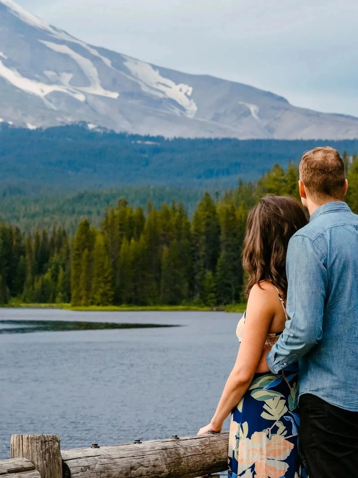 A couple standing on a wooden dock by a lake, surrounded by trees and mountains with snow patches.