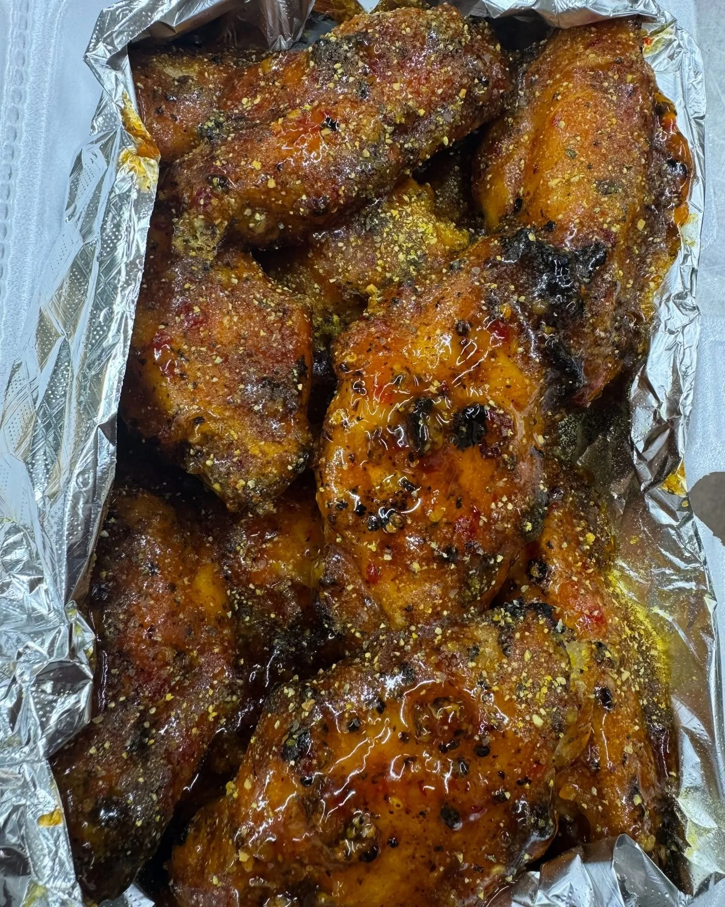 Close-up of spicy glazed chicken drumsticks on aluminum foil, sprinkled with seasoning and black pepper.