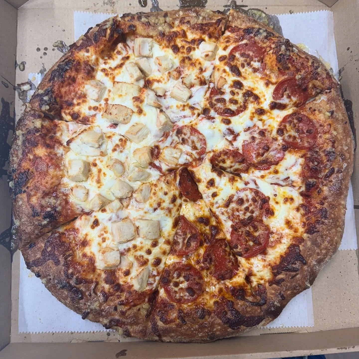 A large pizza in a cardboard takeout box with half topped with chicken and the other half with pepperoni, cheese, and tomato slices.