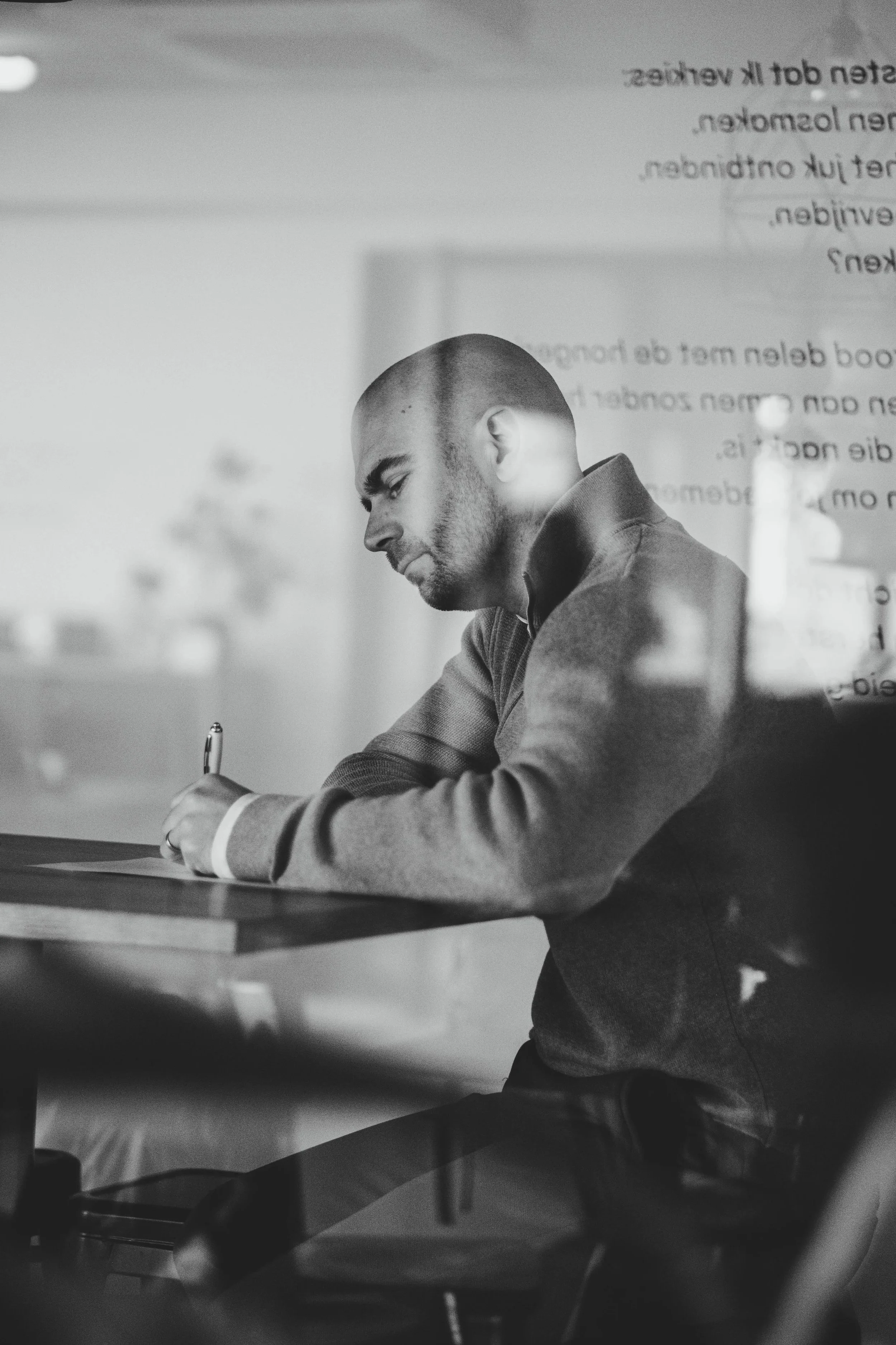 Zwart-wit foto van een man die aan een tafel zit, schriftje schrijft en door een raam kijkt.