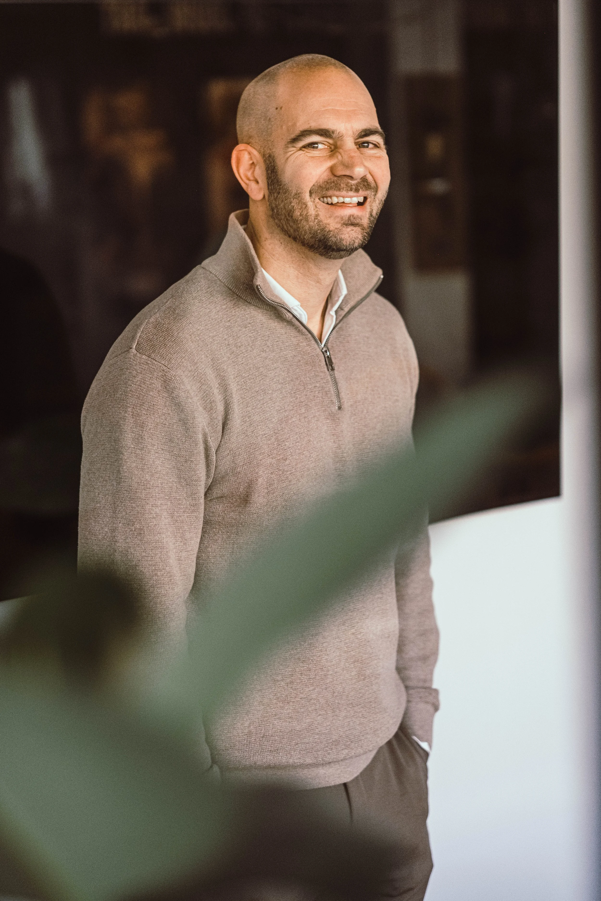 Man met een baard en kale hoofdhuid, lachend, in een lichte grijze trui, binnen.