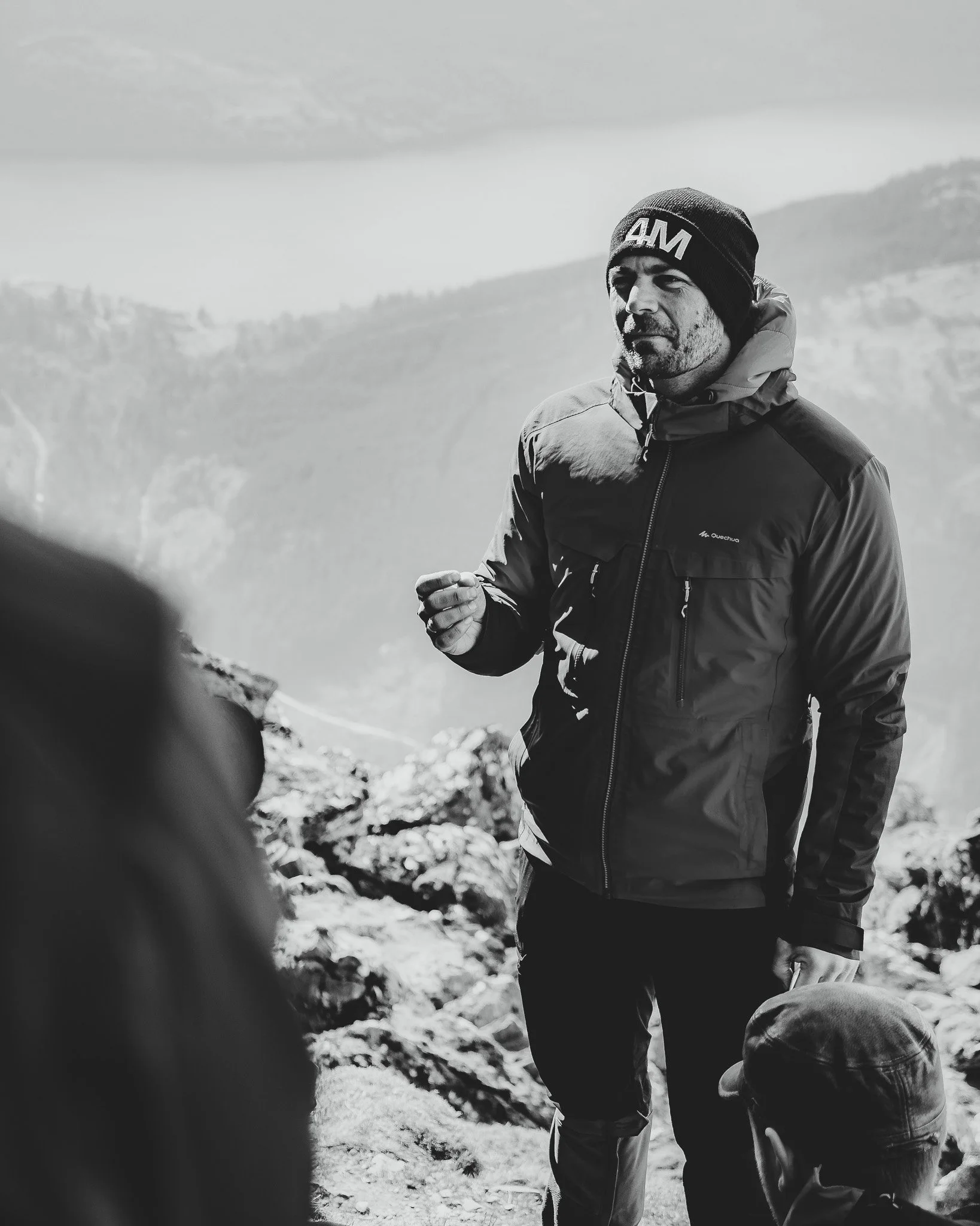 Man in warme kleding en een muts staat op een berghelling, met een berglandschap op de achtergrond, in zwart-wit.