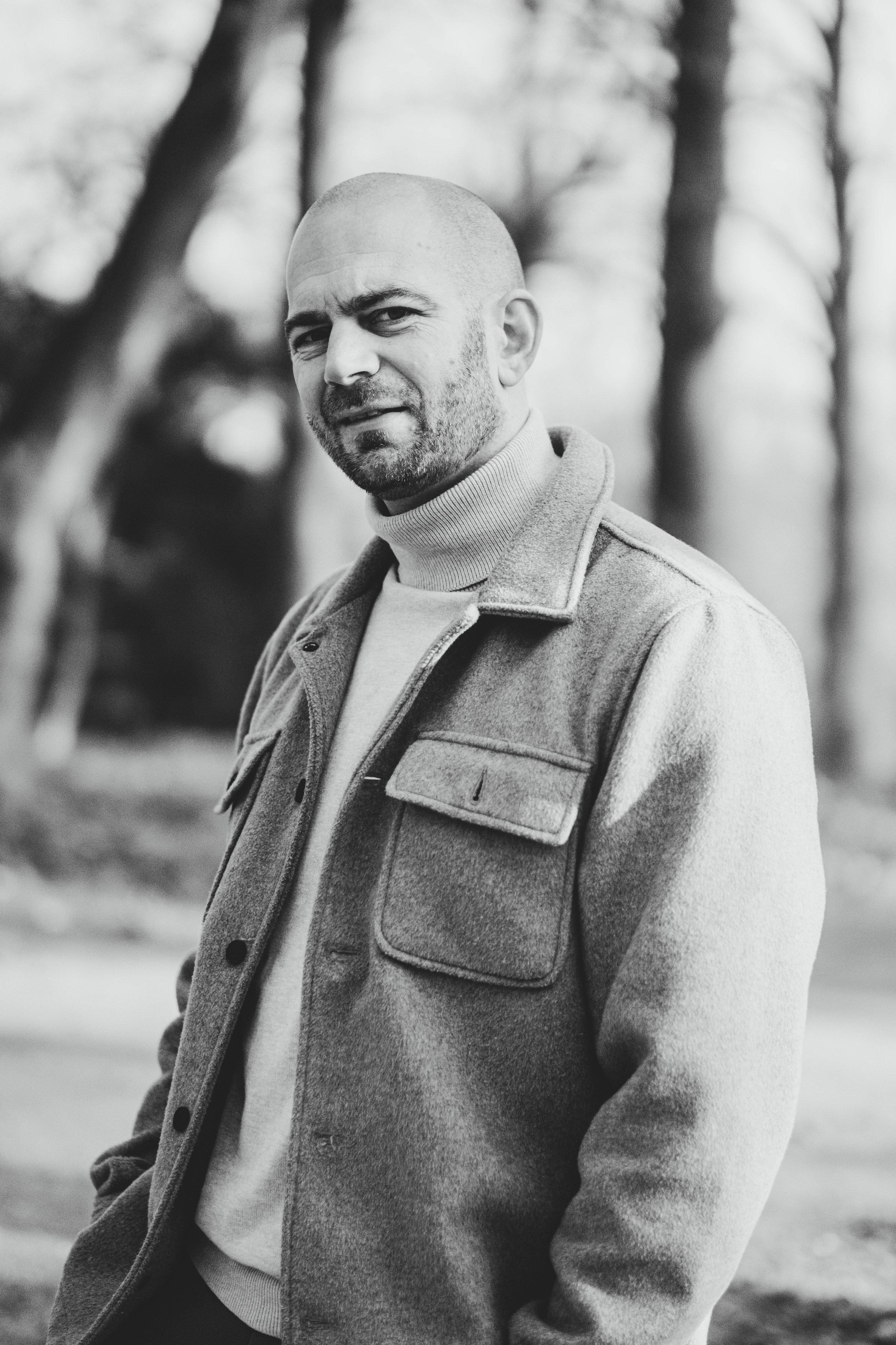 Zwart-wit foto van een man met een kale kop en een baard, gekleed in een jas en een coltrui, staand in een bosrijke omgeving.