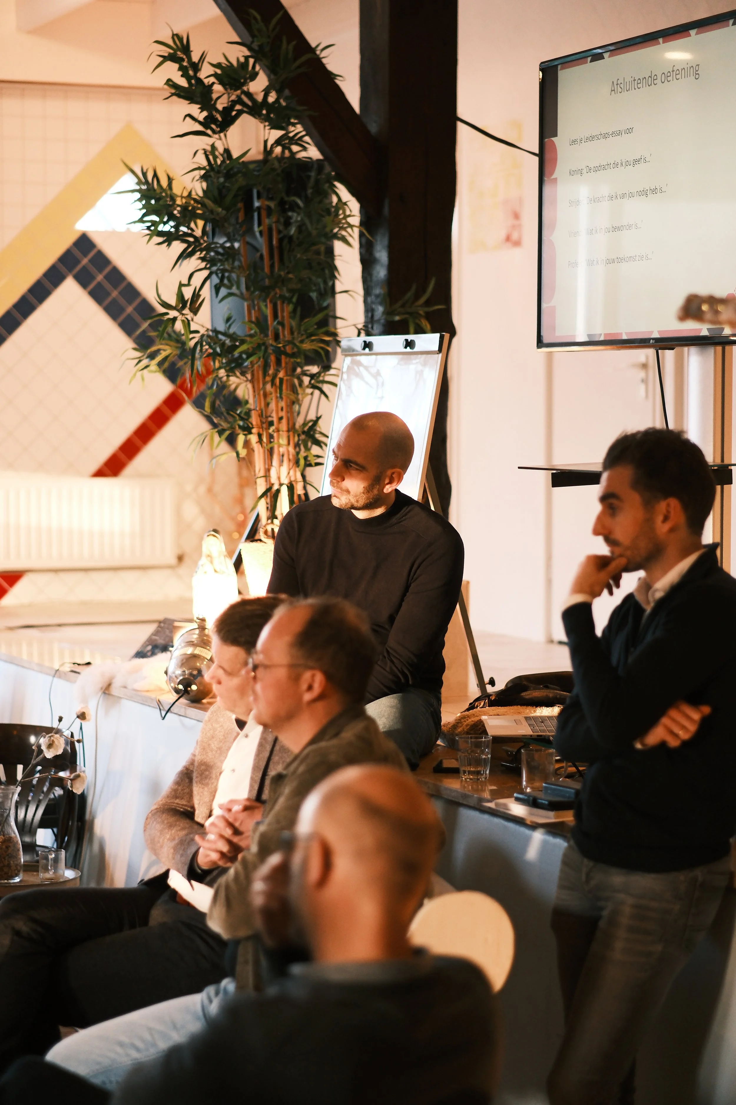 Mensen zitten in een vergaderruimte of conferentiezaal tijdens een presentatie of bijeenkomst, met scherm en flip-over in de achtergrond.