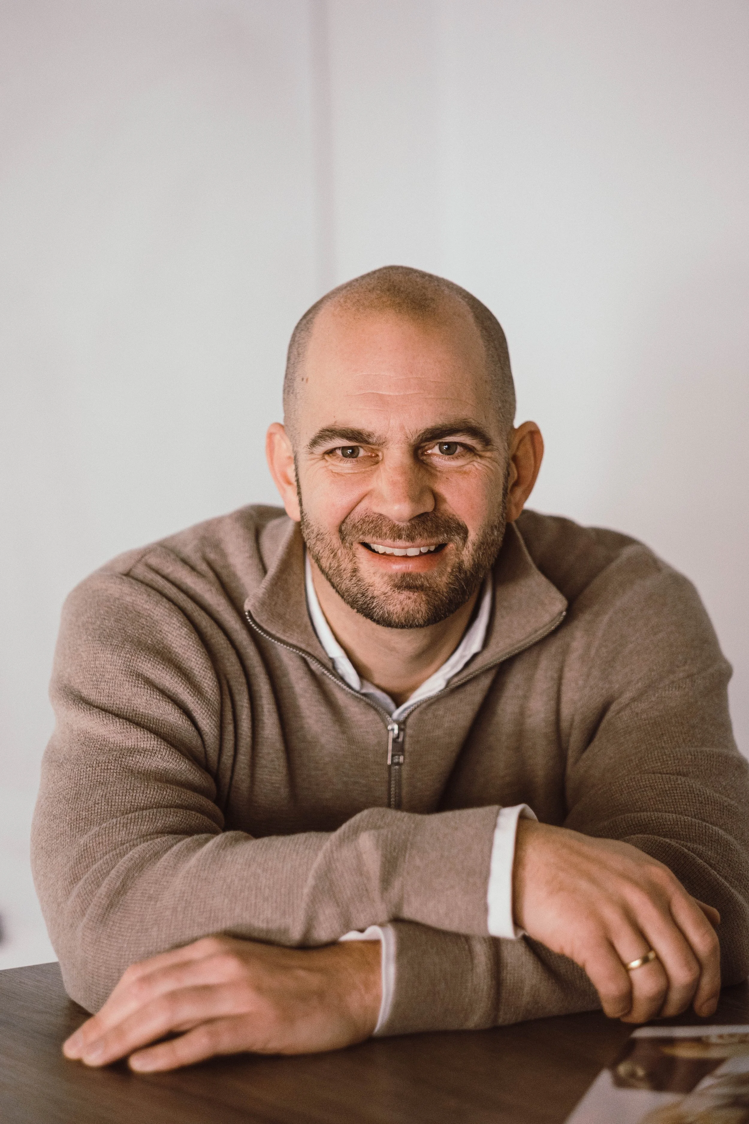 Een glimlachende man met een baard en kale kop, zittend aan een tafel in een nette omgeving.