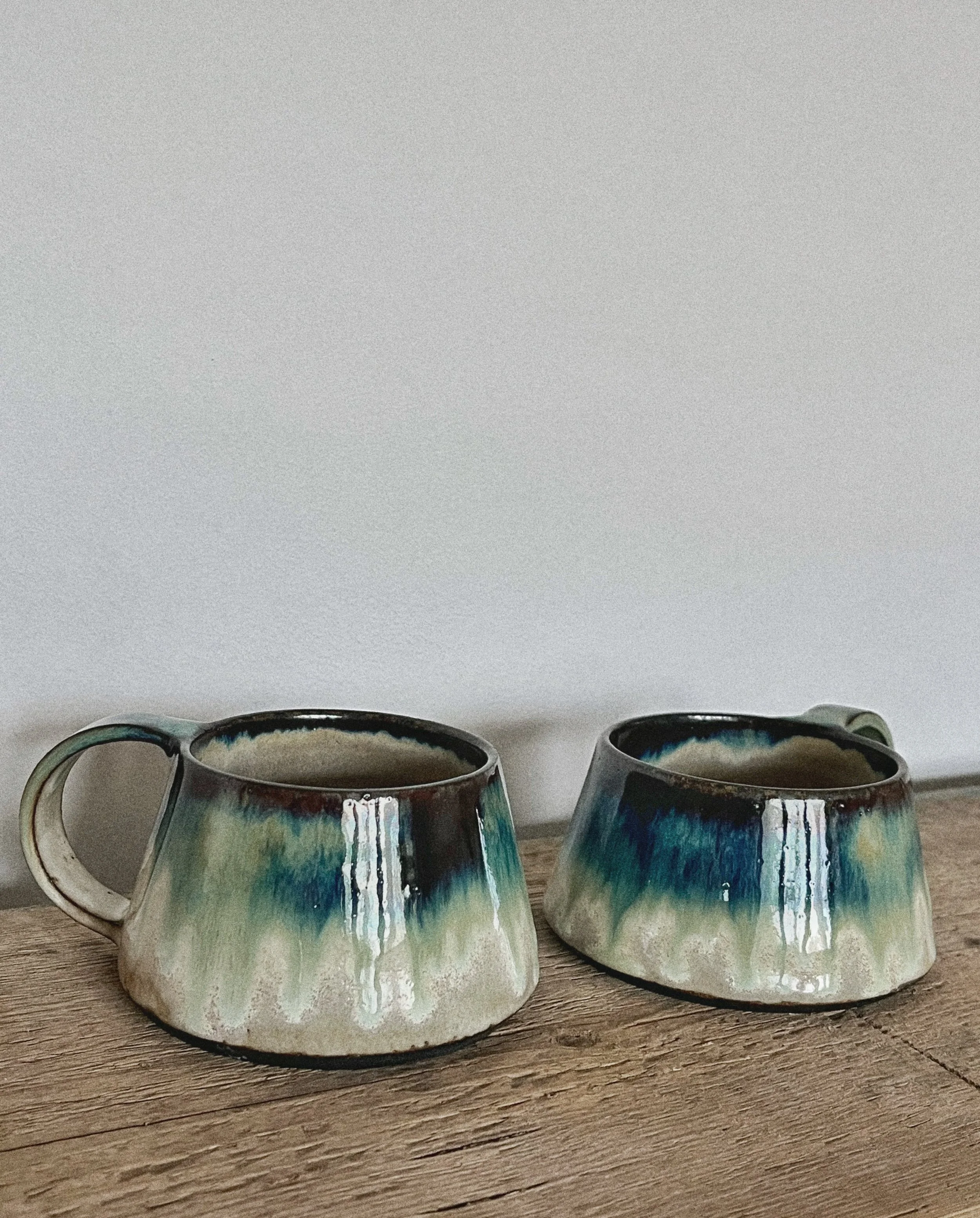Two ceramic coffee mugs with blue, green, and cream glaze sitting on a wooden surface in front of a plain wall.