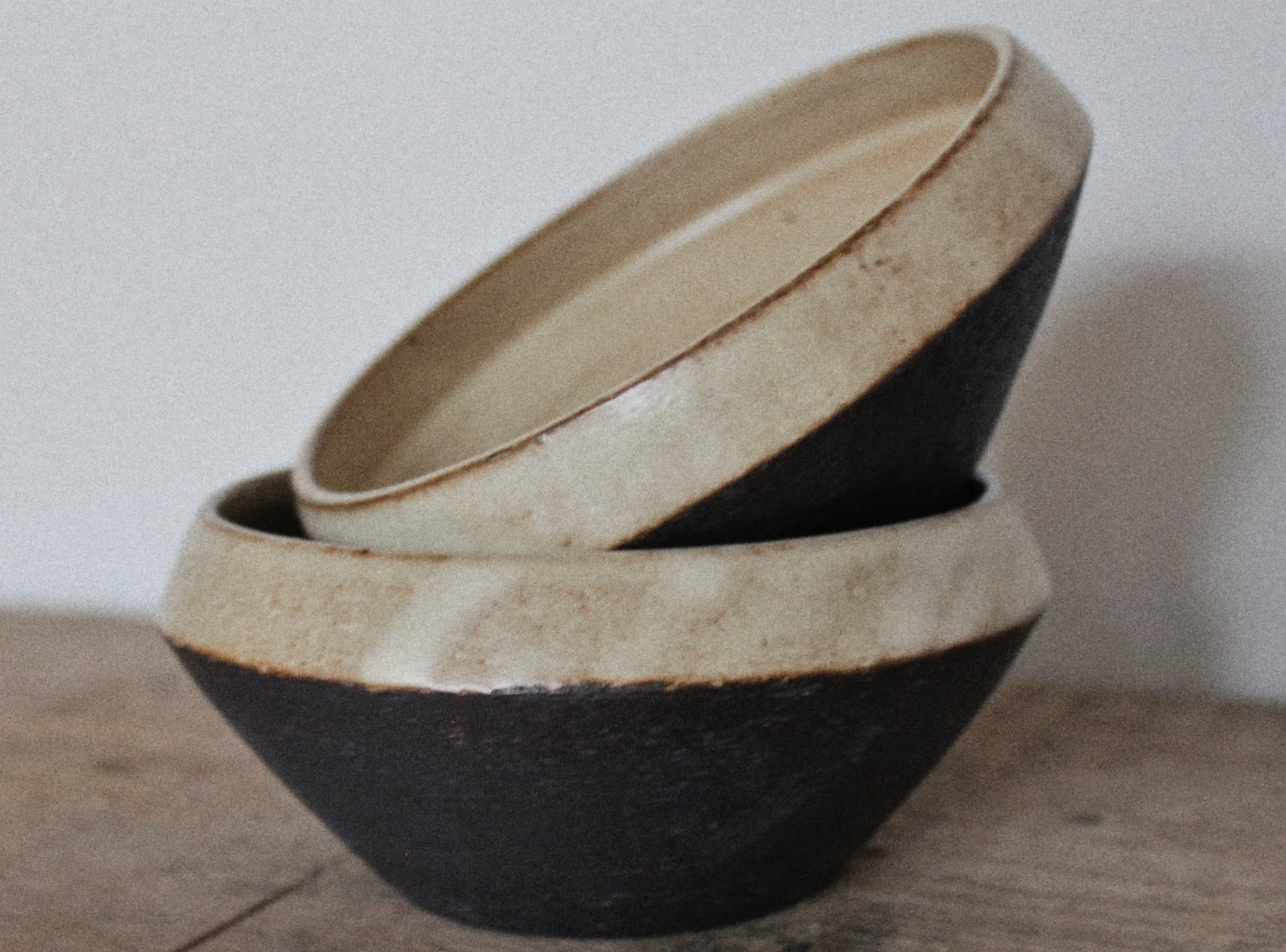 Two stacked bowls, one larger with a black exterior and beige interior, and a smaller inside with a beige exterior and interior, placed on a wooden surface against a plain background.