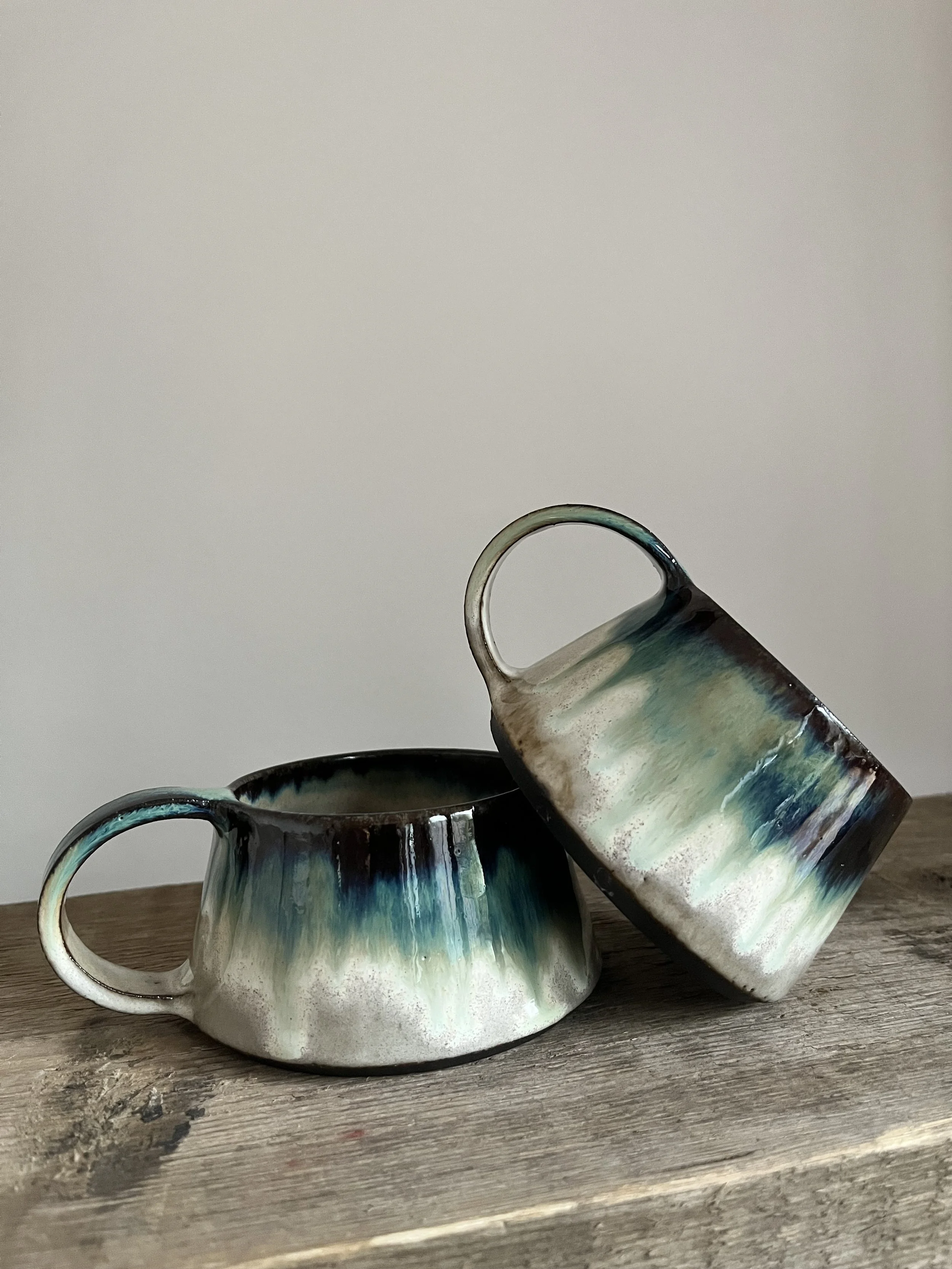 Two ceramic mugs with an abstract blue, green, and brown glaze, one placed inside the other, on a rustic wooden surface against a plain white background.