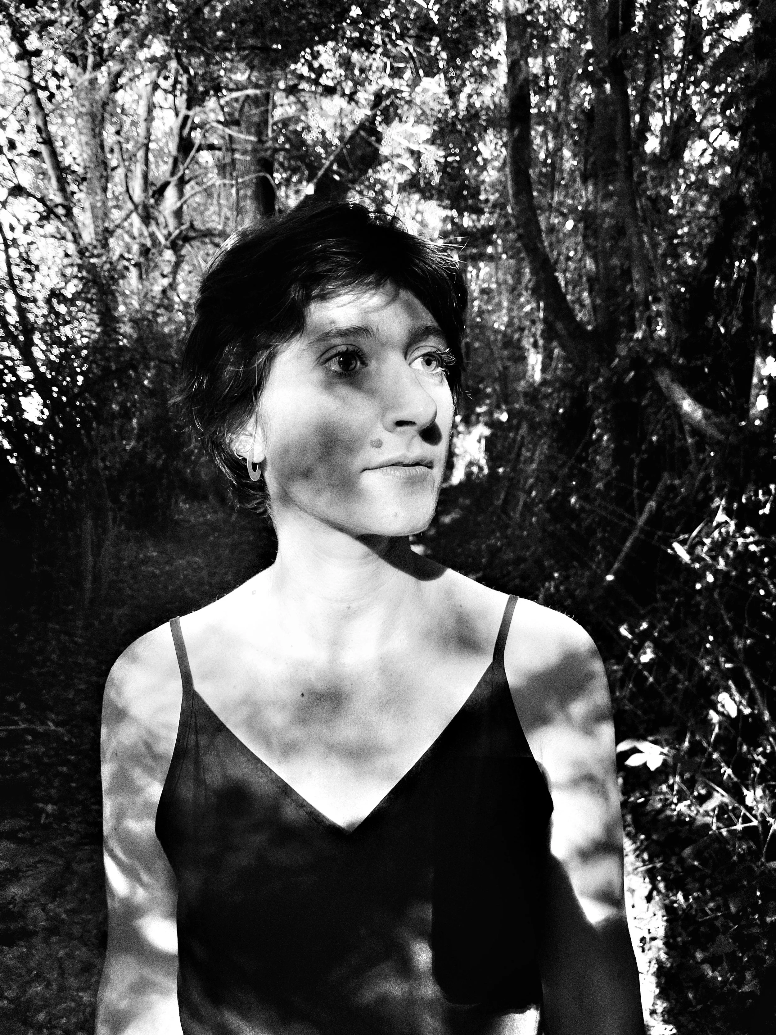A black-and-white photo of a woman with short hair, wearing a sleeveless top, standing in a wooded area with sunlight filtering through the trees.