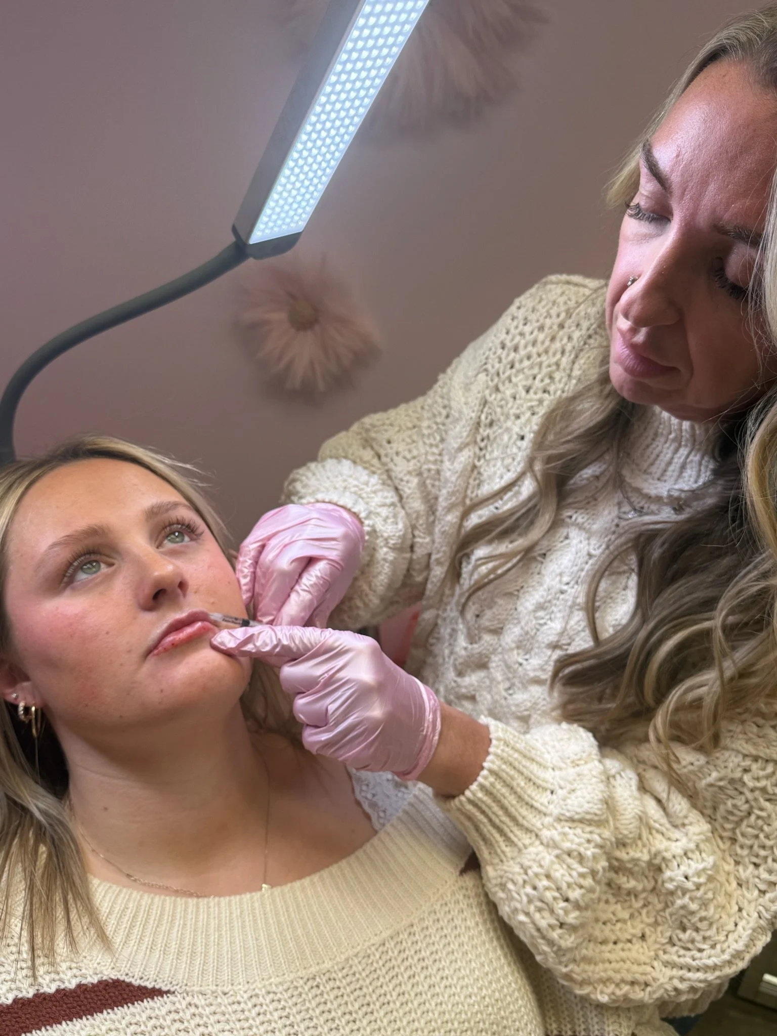 A woman is receiving a cosmetic injection in her lips from a healthcare worker wearing pink gloves, under a bright medical light.