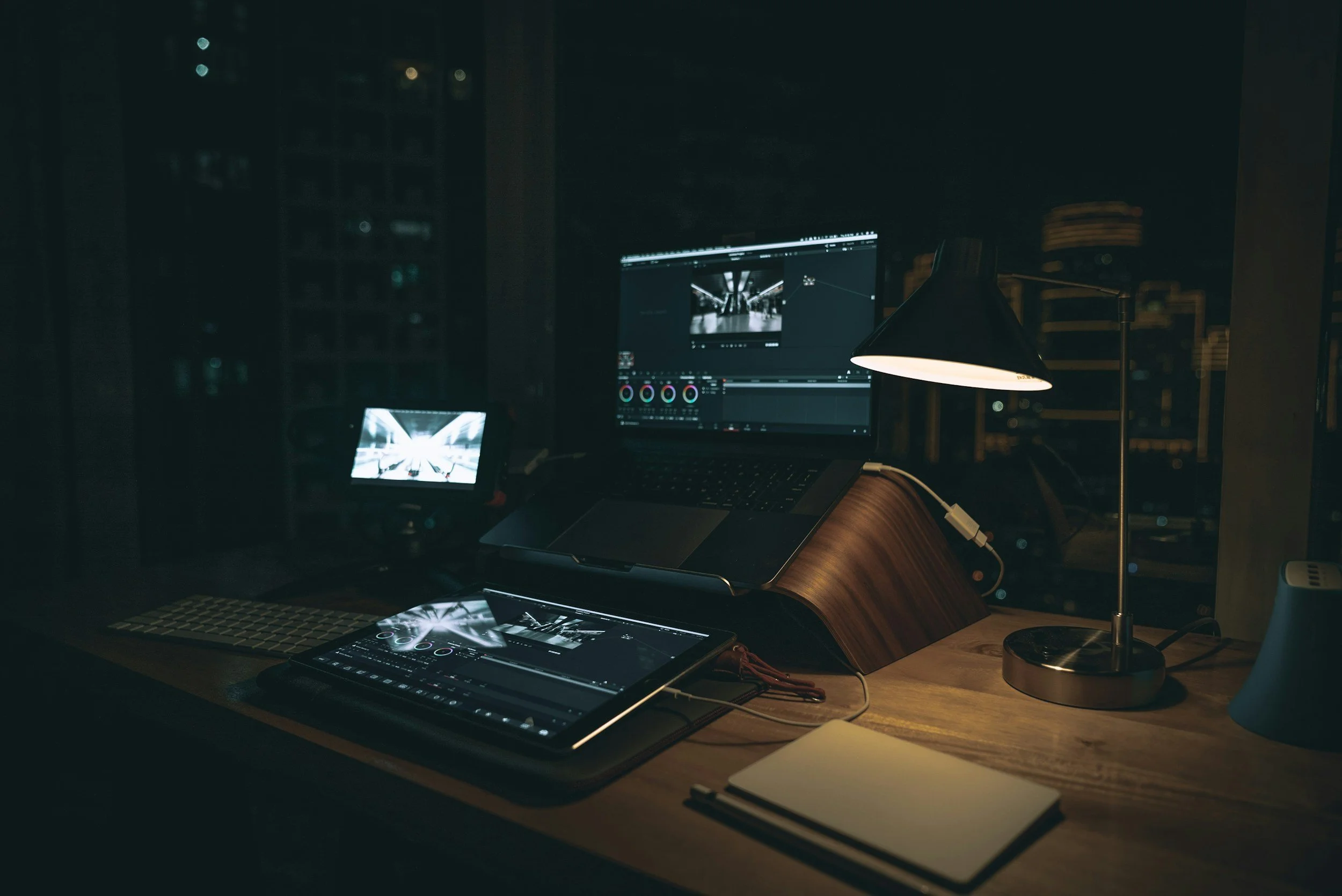 A dimly lit workspace with a laptop, a tablet, and a monitor showing video editing or color grading software. A small lamp provides focused lighting on the wooden desk, which also holds a closed notebook and some cables.