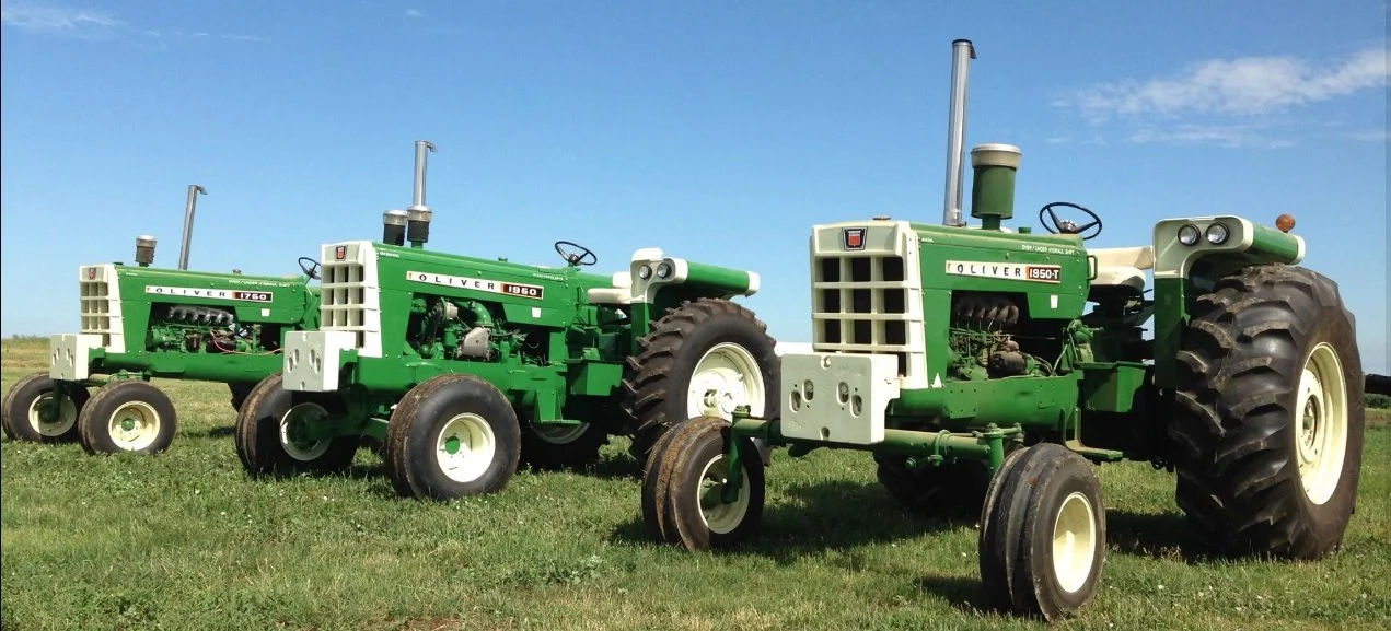 Three vintage green and white Oliver tractors of different sizes parked on grass field under blue sky.