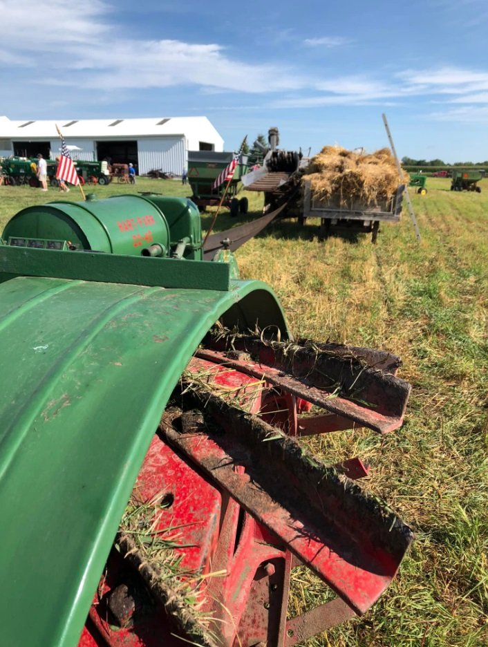 Farm equipment with green and red machinery, hay on a trailer, and a large white building in the background on a sunny day.