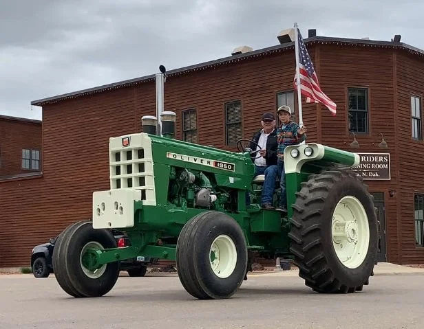 Two children sitting on a green Oliver tractor with an American flag attached, parked in front of a brown historic building with a sign reading "Riders Meeting Room."