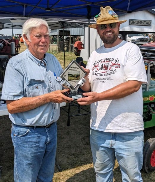 An elderly man and a younger man are outdoors under a blue canopy, shaking hands while exchanging a trophy. The elderly man wears a light blue shirt and jeans, and the younger man wears a straw hat, sunglasses, a white t-shirt with red print, and jeans. There are tents, equipment, and vehicles in the background.