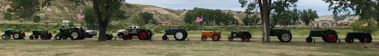 A lineup of vintage tractors and farm equipment on a grassy area with trees and hills in the background, some with American flags.