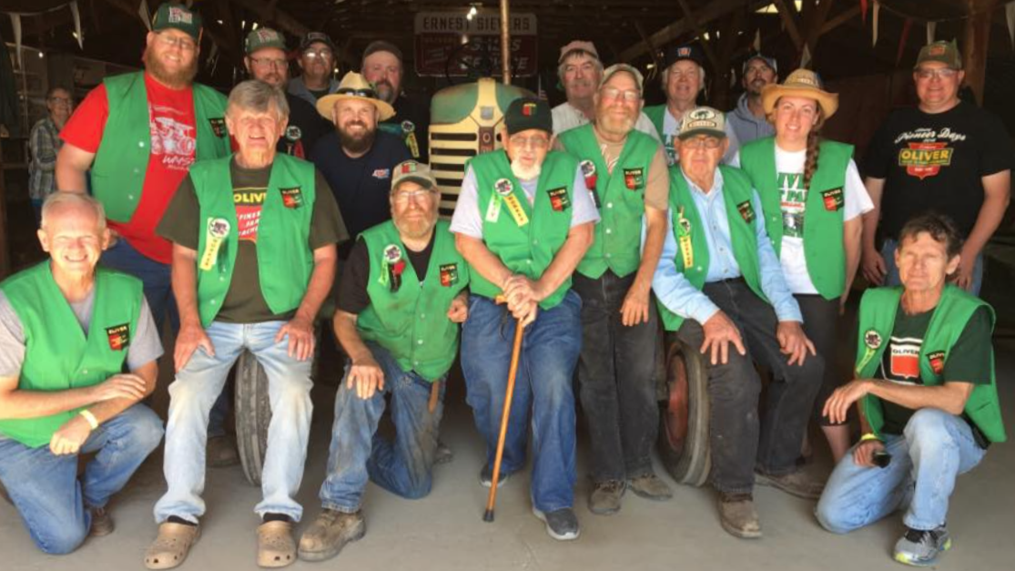 Group of people, mostly elderly, wearing green vests with patches, gathered in a rustic indoor setting, some wearing hats, some kneeling and some standing, smiling at the camera.