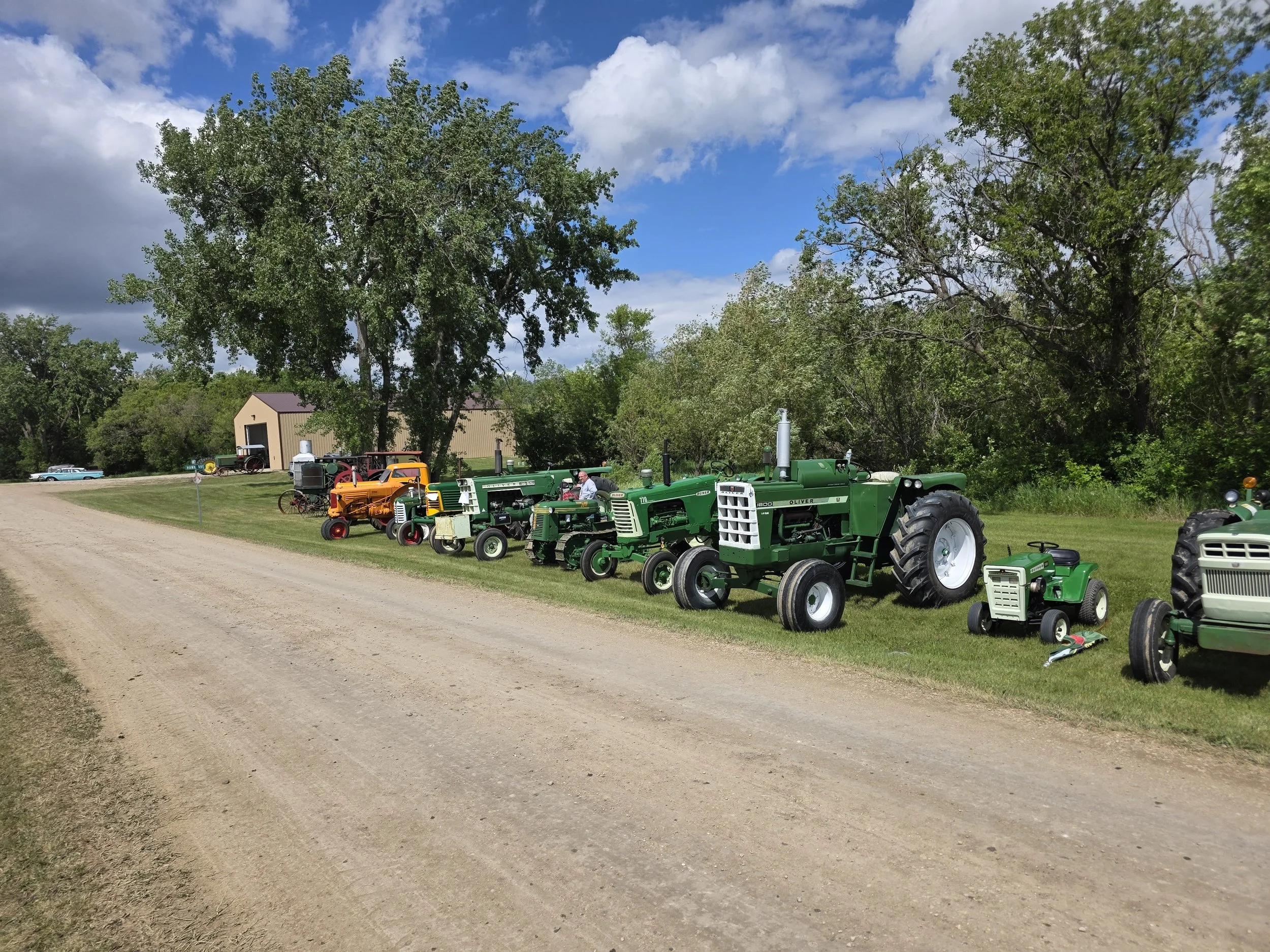 A display of vintage tractors lined up along a grassy area on a farm, with trees and a barn in the background under a partly cloudy sky.