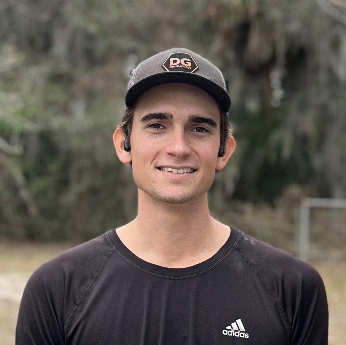 A young man with dark hair, wearing a gray and black cap with a logo, black Adidas t-shirt, and wireless earbuds, smiling outdoors with blurred trees in the background.