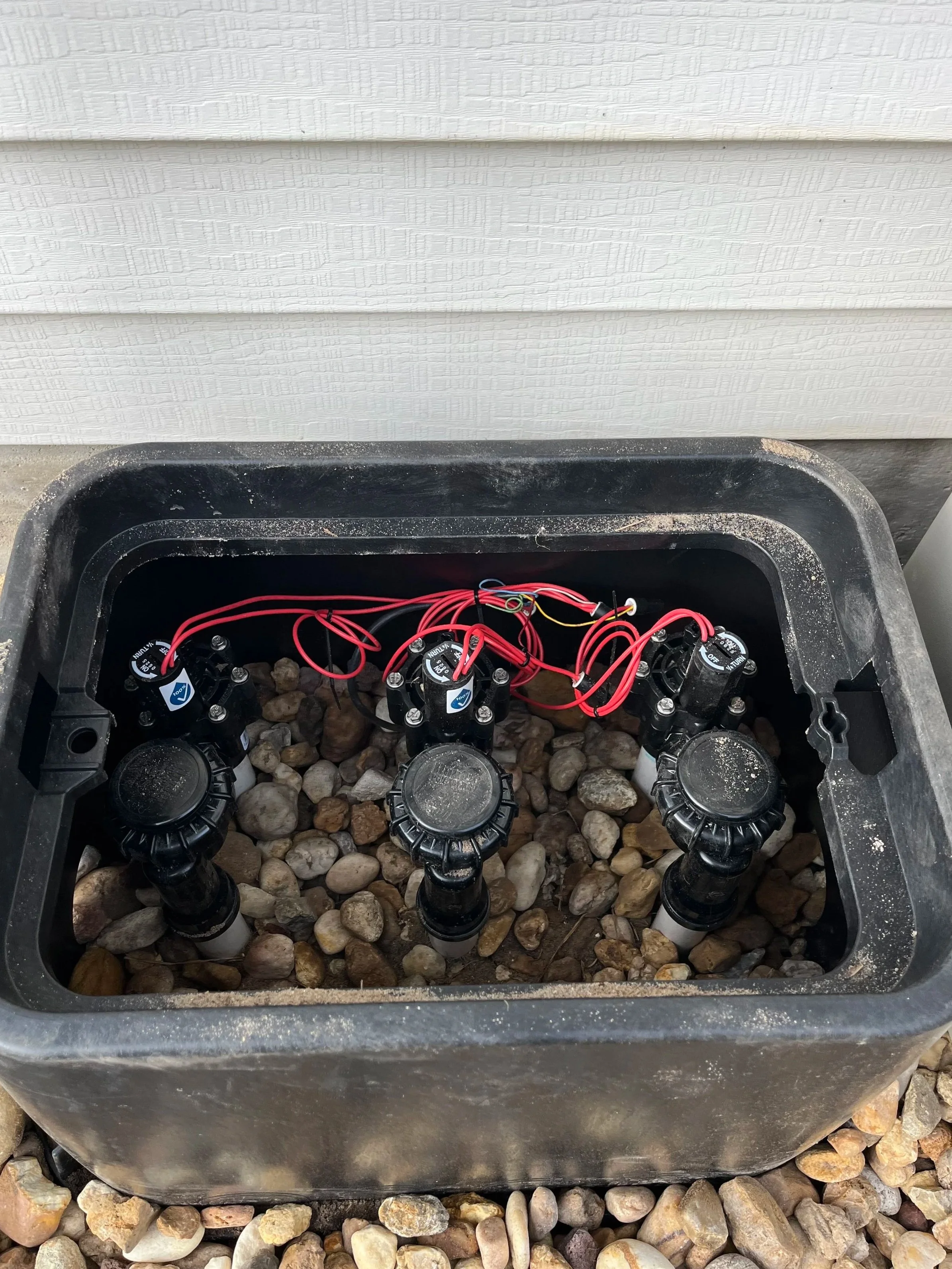 A black box with three sprinkler valves and red wires inside, placed on gravel in front of a white wall.