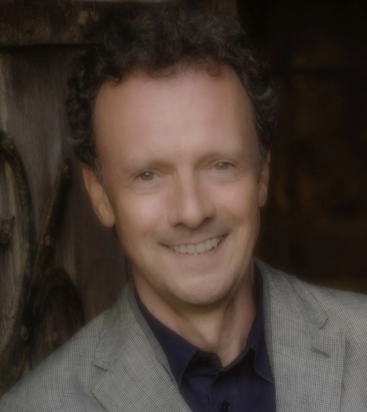 A man with curly dark hair, light skin, and a friendly smile wearing a gray blazer and a dark shirt, standing outdoors against a wooden background.