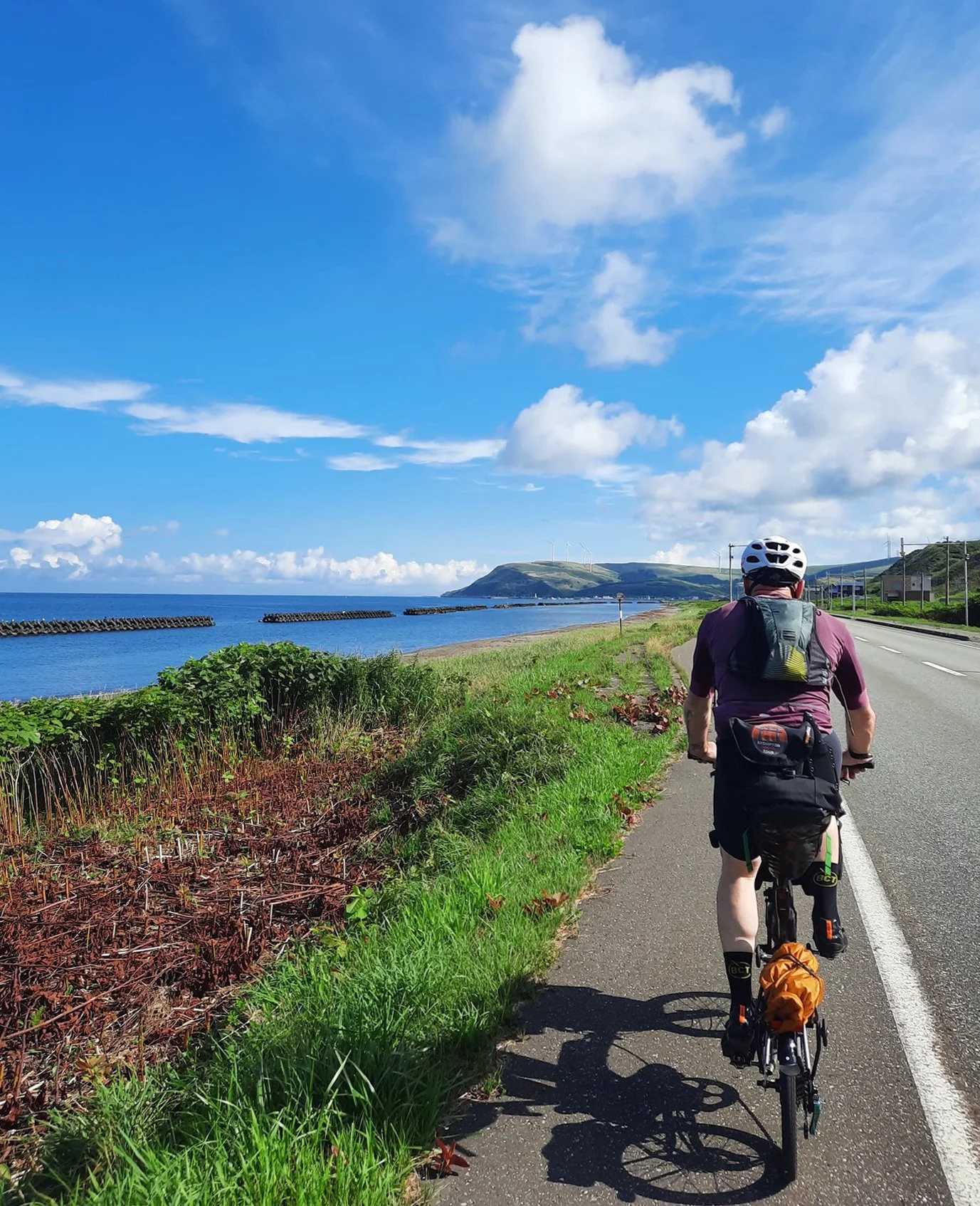 Person riding a bicycle along a coastal road with green grass, blue sky, and clouds, with a view of the ocean and hills in the background.