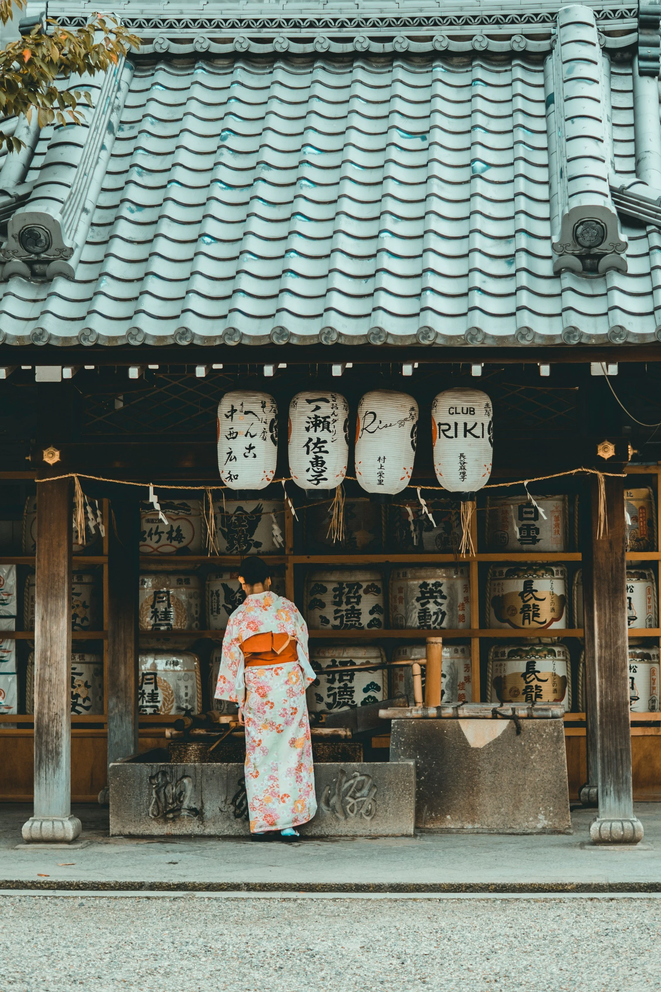 A woman in a floral kimono standing at a traditional Japanese temple water basin surrounded by lanterns and wooden structures.