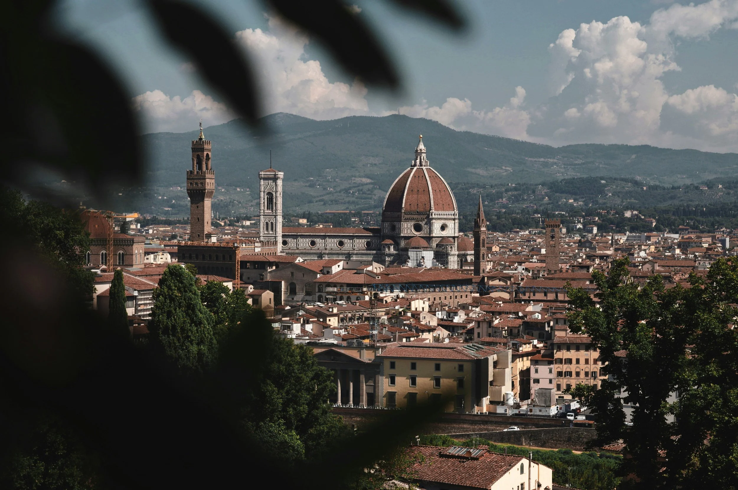 View of Florence, Italy, featuring the Florence Cathedral with its red-tiled dome, surrounded by historic buildings and green trees, with hills in the background under a partly cloudy sky.