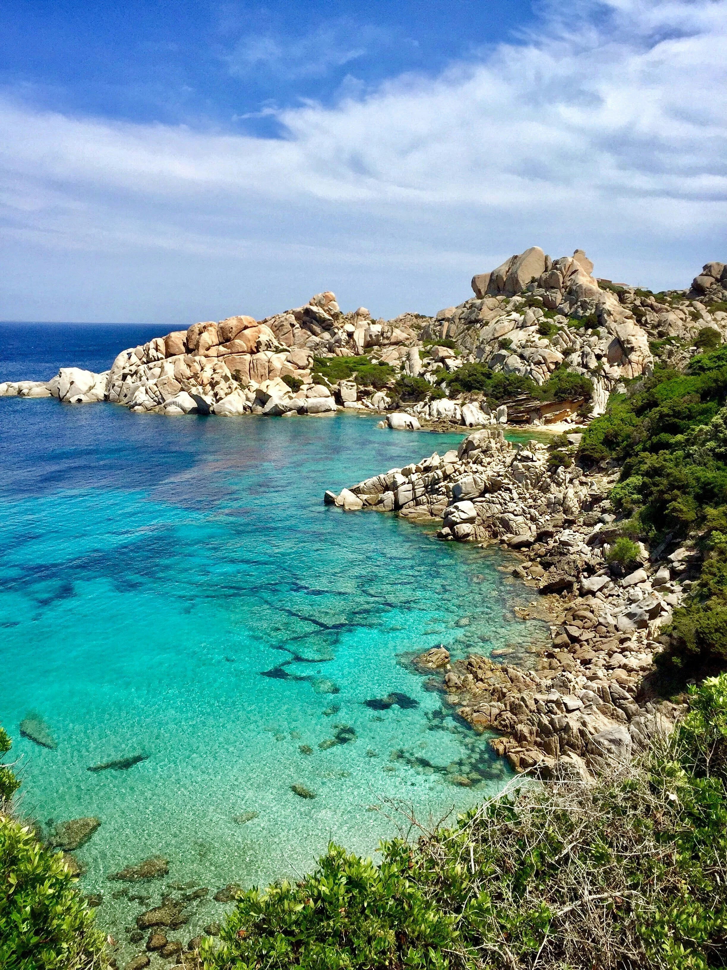 Scenic coastal landscape with turquoise water, rocky shoreline, and green shrubbery under a partly cloudy sky.