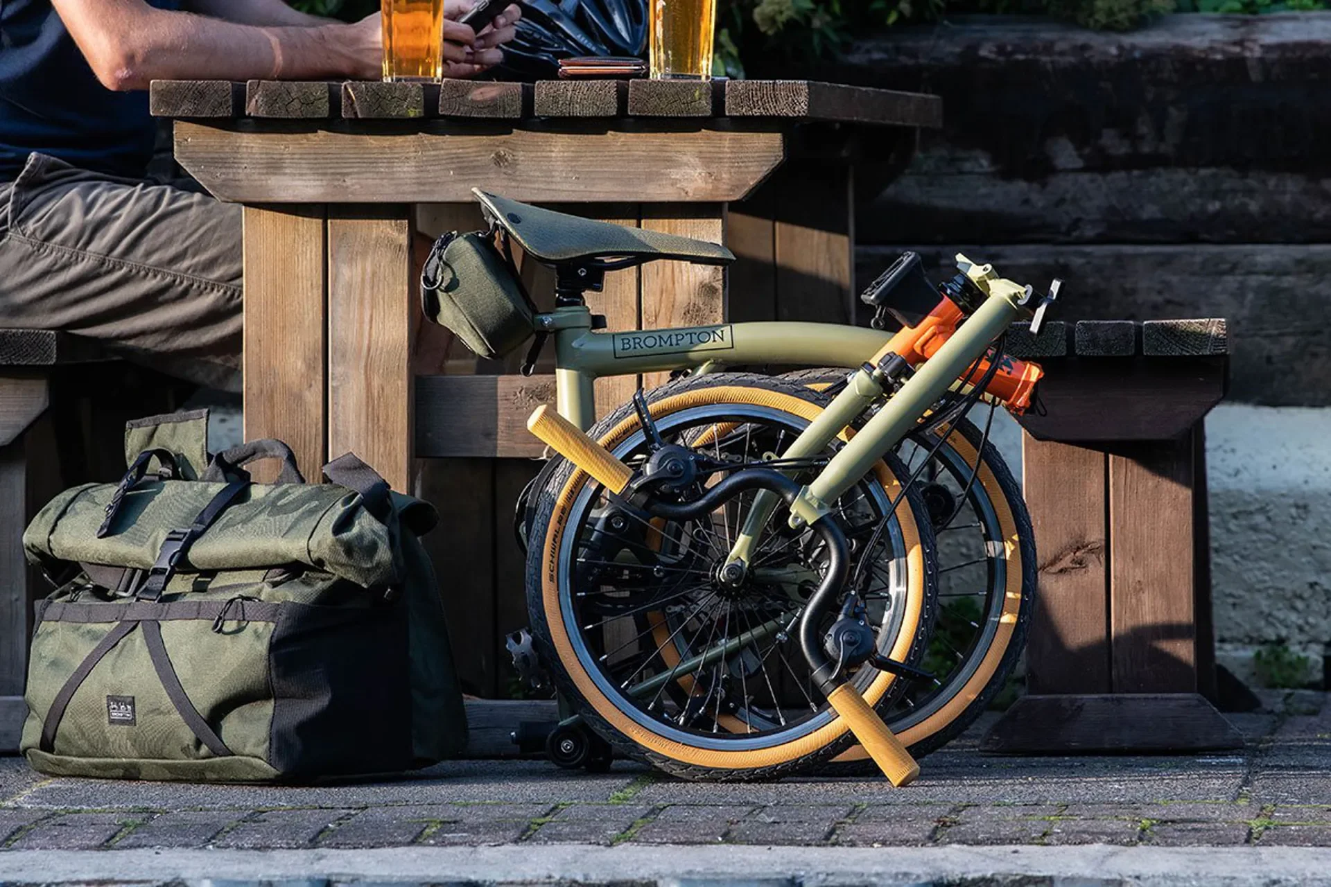 A folded green bicycle with orange tires leaning against a wooden picnic table. A green and black backpack is on the ground nearby. Two glasses of beer are on the table, and a person is sitting at the table using a smartphone.
