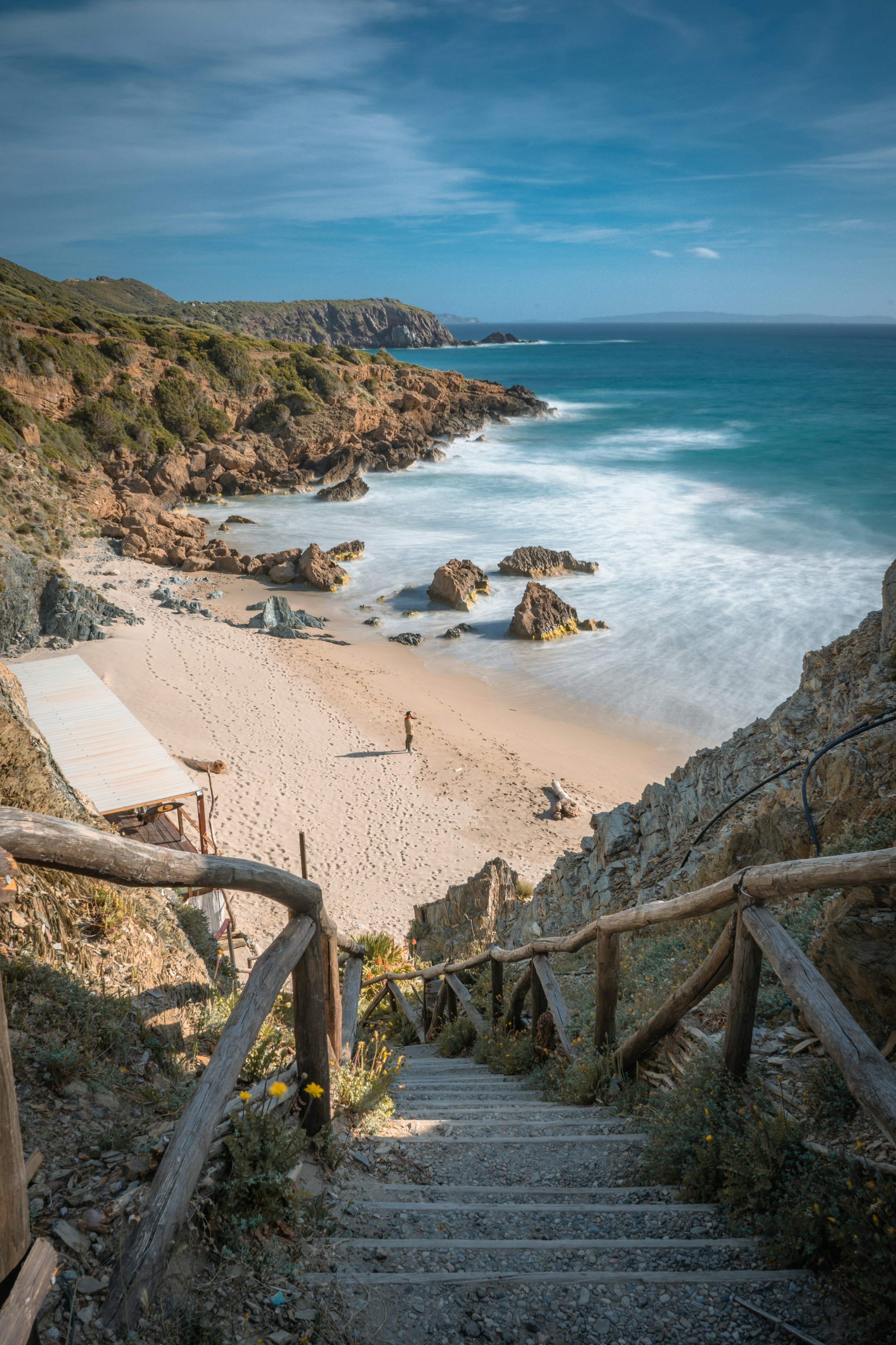 A staircase made of wood and stone leads down to a sandy beach with rocks and cliffs on the coast, under a blue sky.