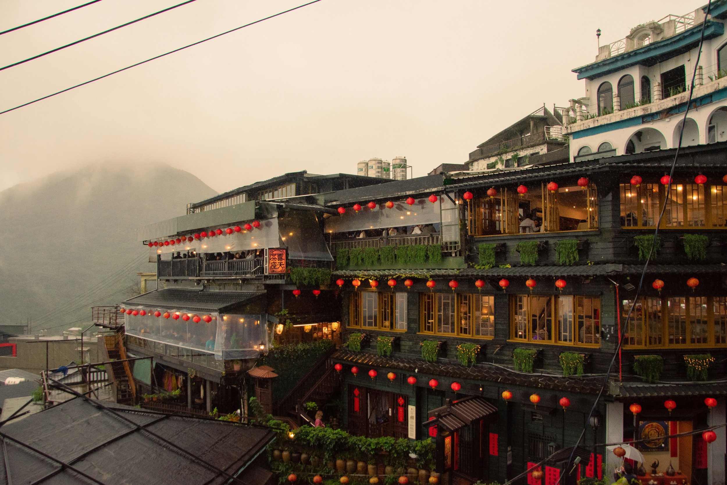 Multi-story traditional Asian-style building decorated with numerous red lanterns, situated on a hillside with greenery and neighboring buildings, during evening.